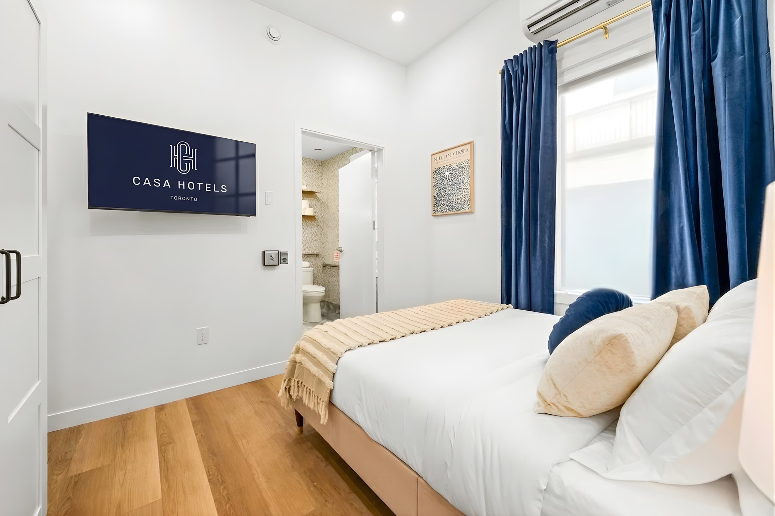 Bedroom with a beige upholstered bed, light wood flooring, a wall-mounted TV displaying "CASA HOTELS TORONTO" logo, and an open doorway leading to a bathroom.