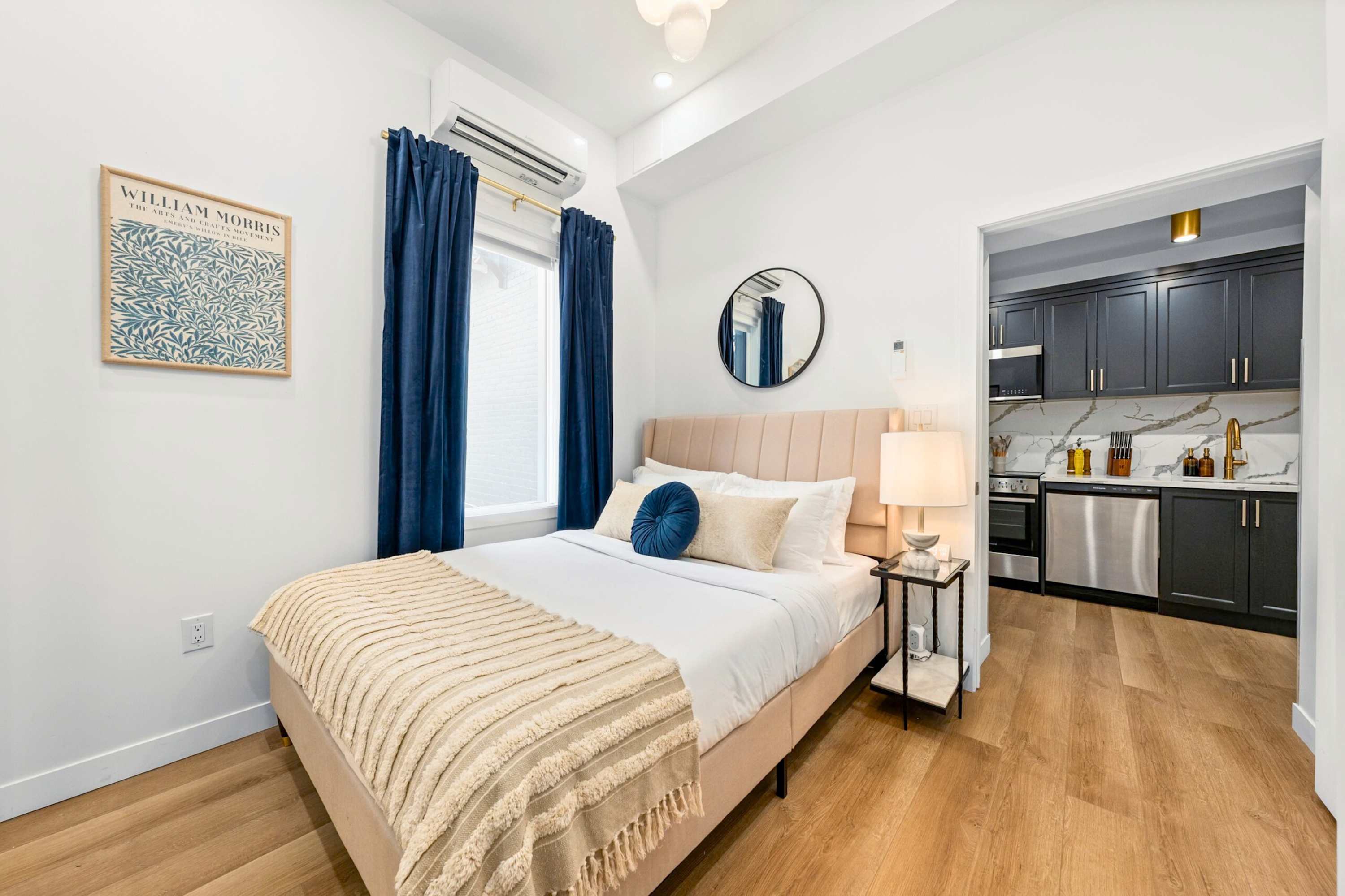 Bright bedroom with light wood flooring, a beige upholstered bed with blue and cream pillows, blue curtains, and a view into the adjacent modern kitchen.