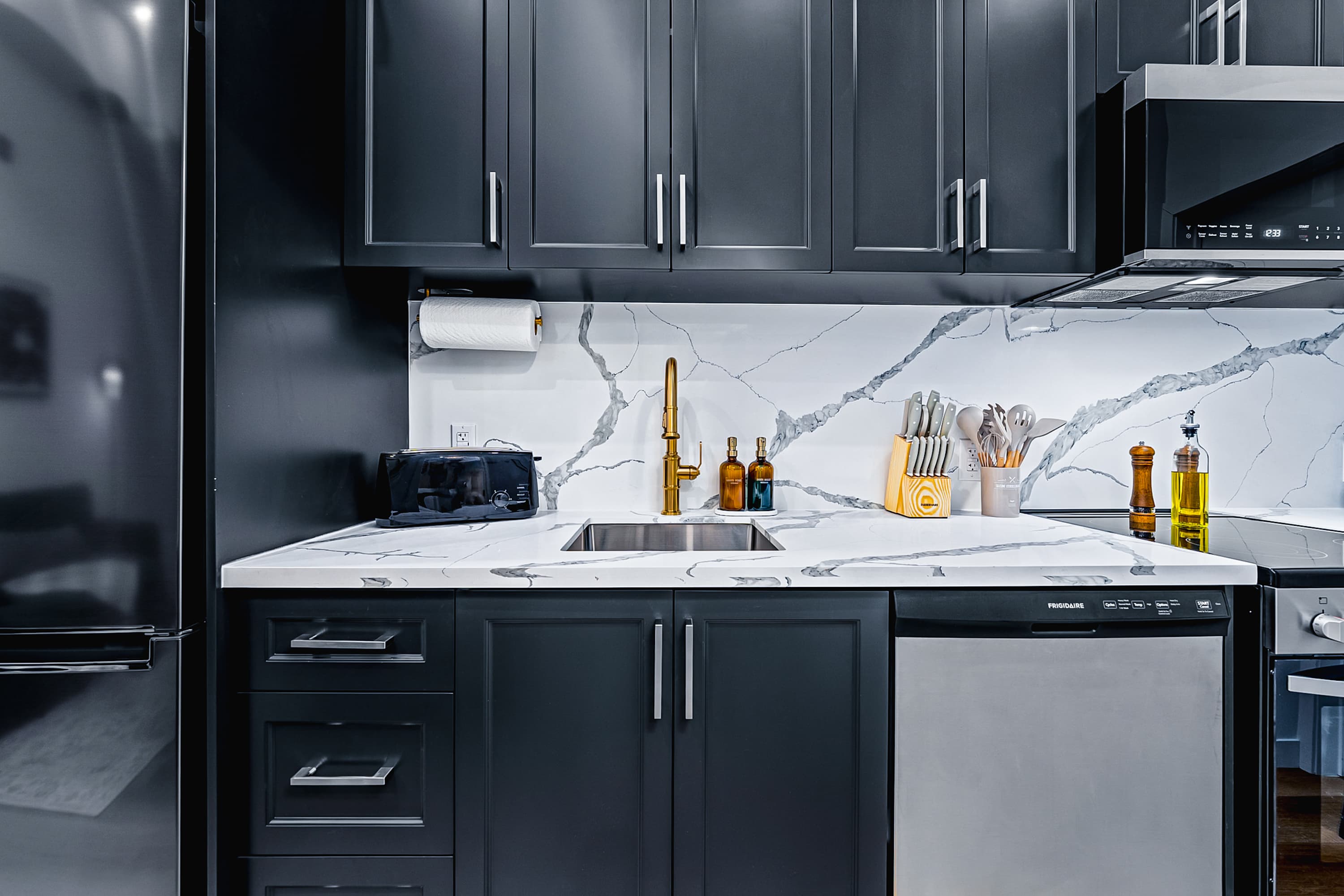 A close-up of a modern kitchen with dark grey shaker-style cabinets, white marble-patterned countertops and backsplash, a gold gooseneck faucet, and stainless steel appliances including a dishwasher and refrigerator.