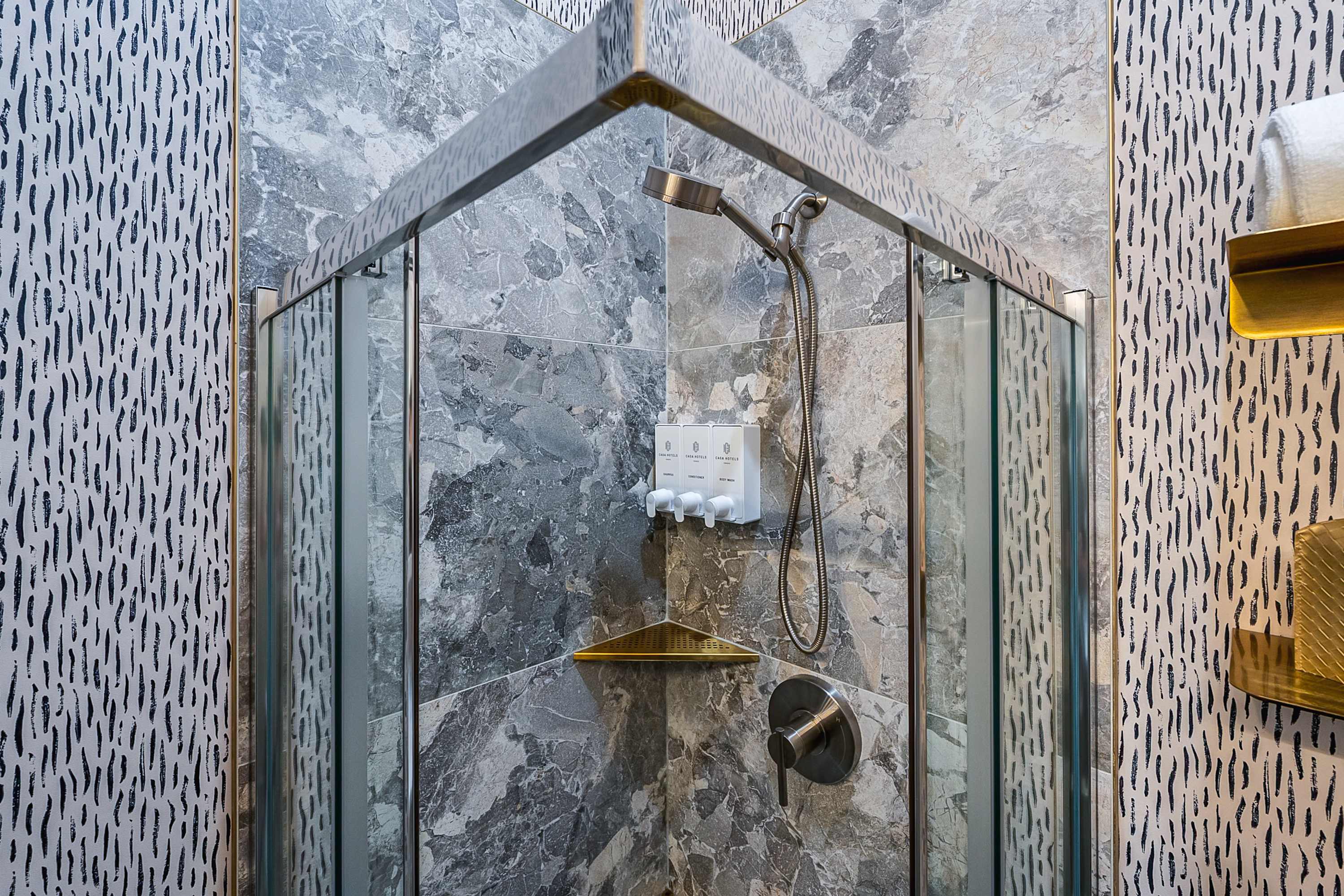 A close-up view inside the glass-enclosed shower, showcasing grey marble-patterned tiles, a handheld showerhead, a corner shelf, and wall-mounted dispensers for toiletries.