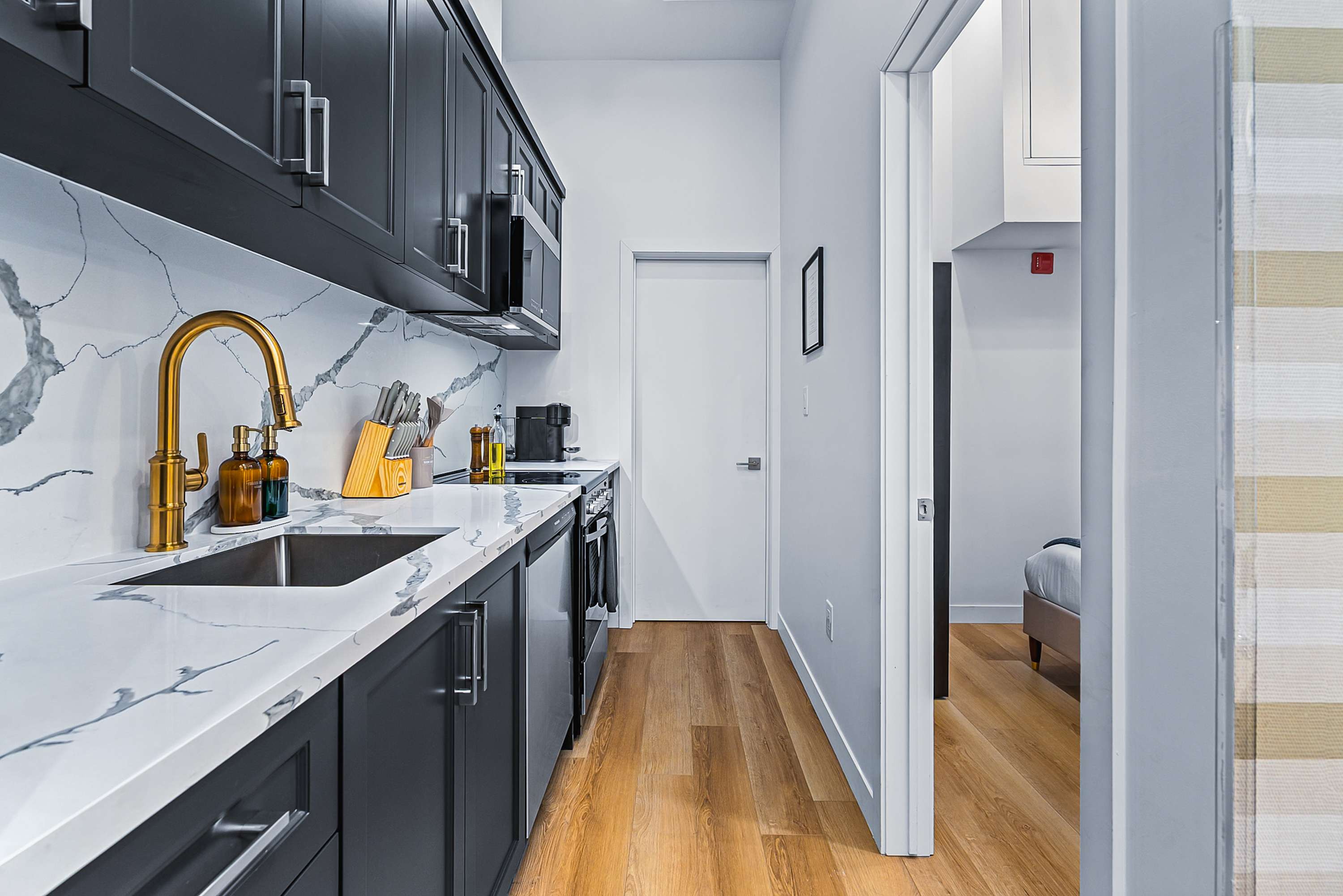 A narrow, modern kitchen with dark grey cabinetry, white marble-patterned countertops and backsplash, a gold-toned faucet, and stainless steel appliances, leading to a bedroom.