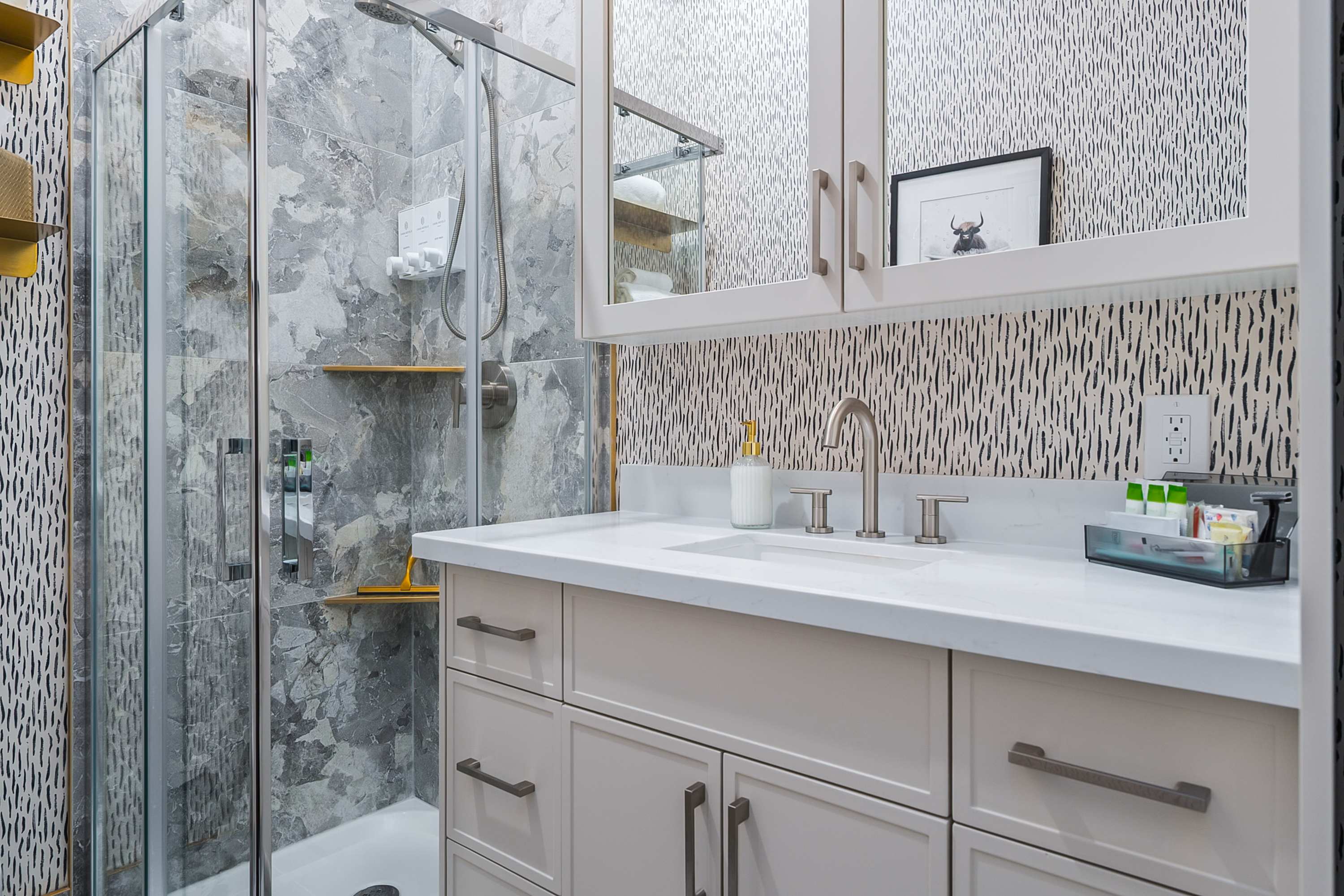 A modern bathroom with a white vanity featuring a single sink, brushed nickel faucet, and white quartz countertop, adjacent to a glass-enclosed shower with grey marble-patterned tiles and a corner shelf.