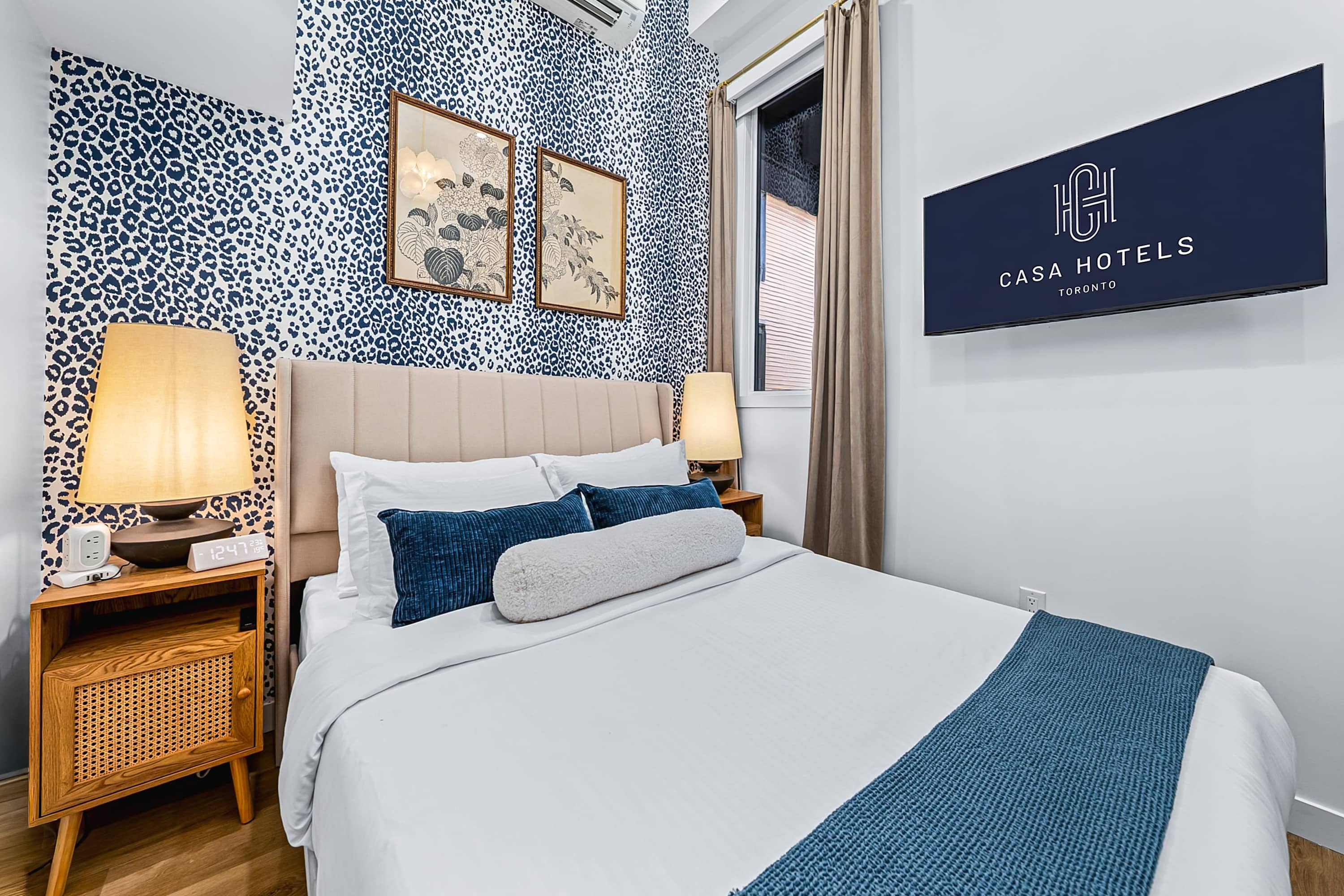 A cozy bedroom with a queen-sized bed featuring a beige upholstered headboard, blue and white bedding, and a striking blue leopard print wallpaper accent wall, with a wall-mounted TV.