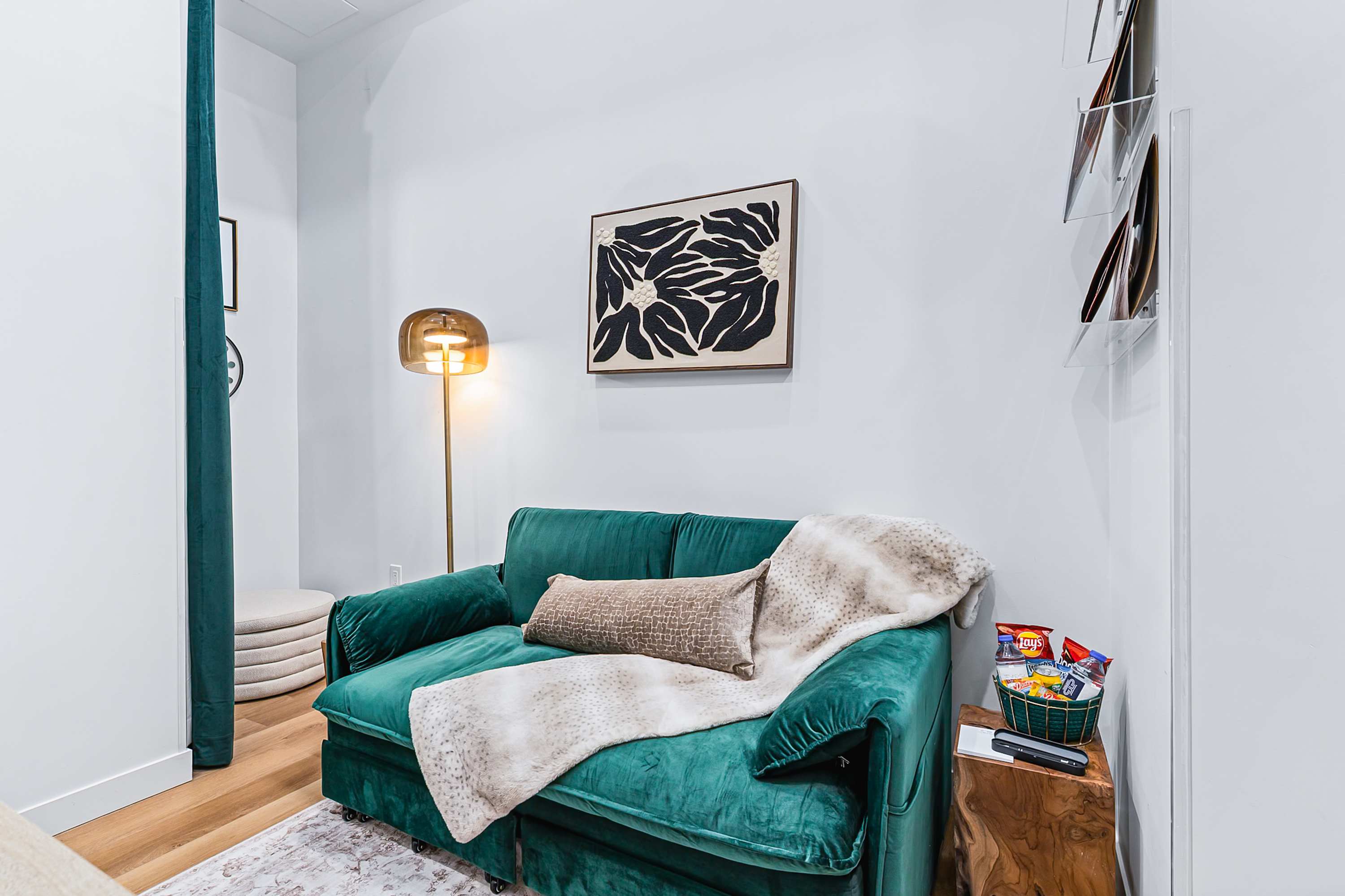 A plush emerald green sofa adorned with a faux fur throw and decorative pillows, positioned next to a rustic wooden side table holding snacks, with a modern floor lamp and abstract art on the wall.