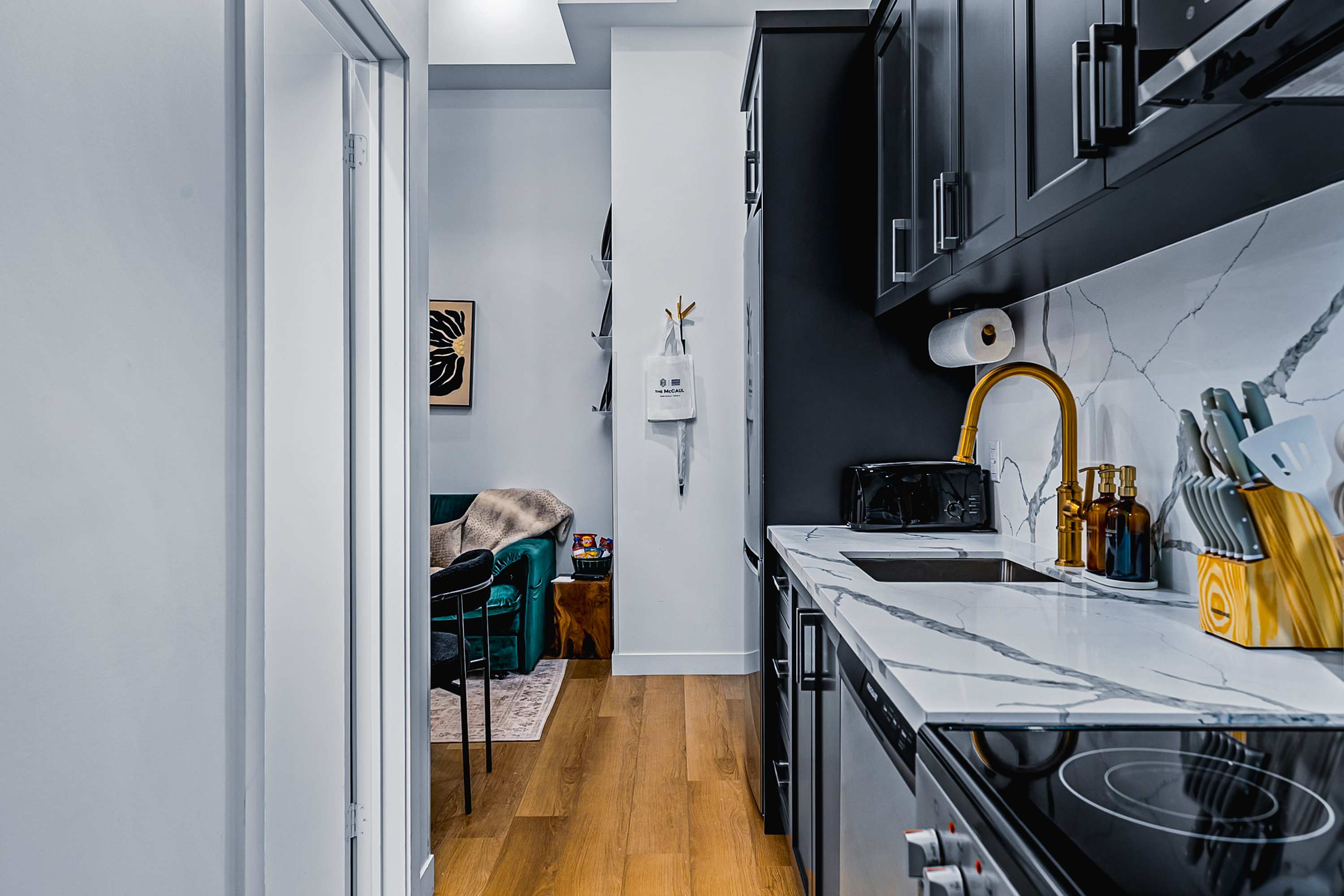A modern galley kitchen with dark grey cabinets, white marble-patterned countertops, a gold faucet, and an electric stove, opening into a living area with a green velvet couch.