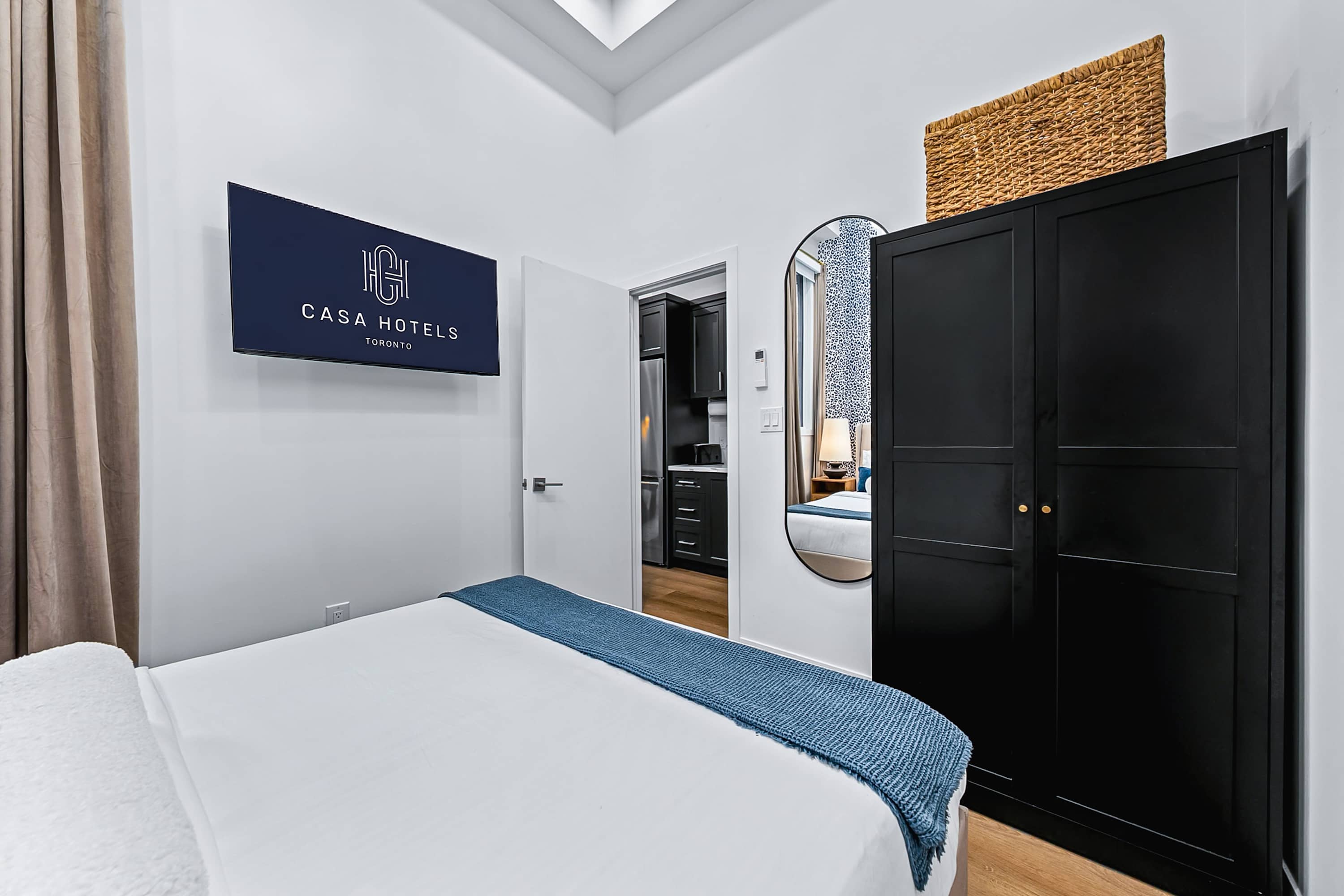 A view of bedroom 1 with a queen bed, a black wardrobe, and a large oval mirror, showing the open doorway leading to the kitchen area.