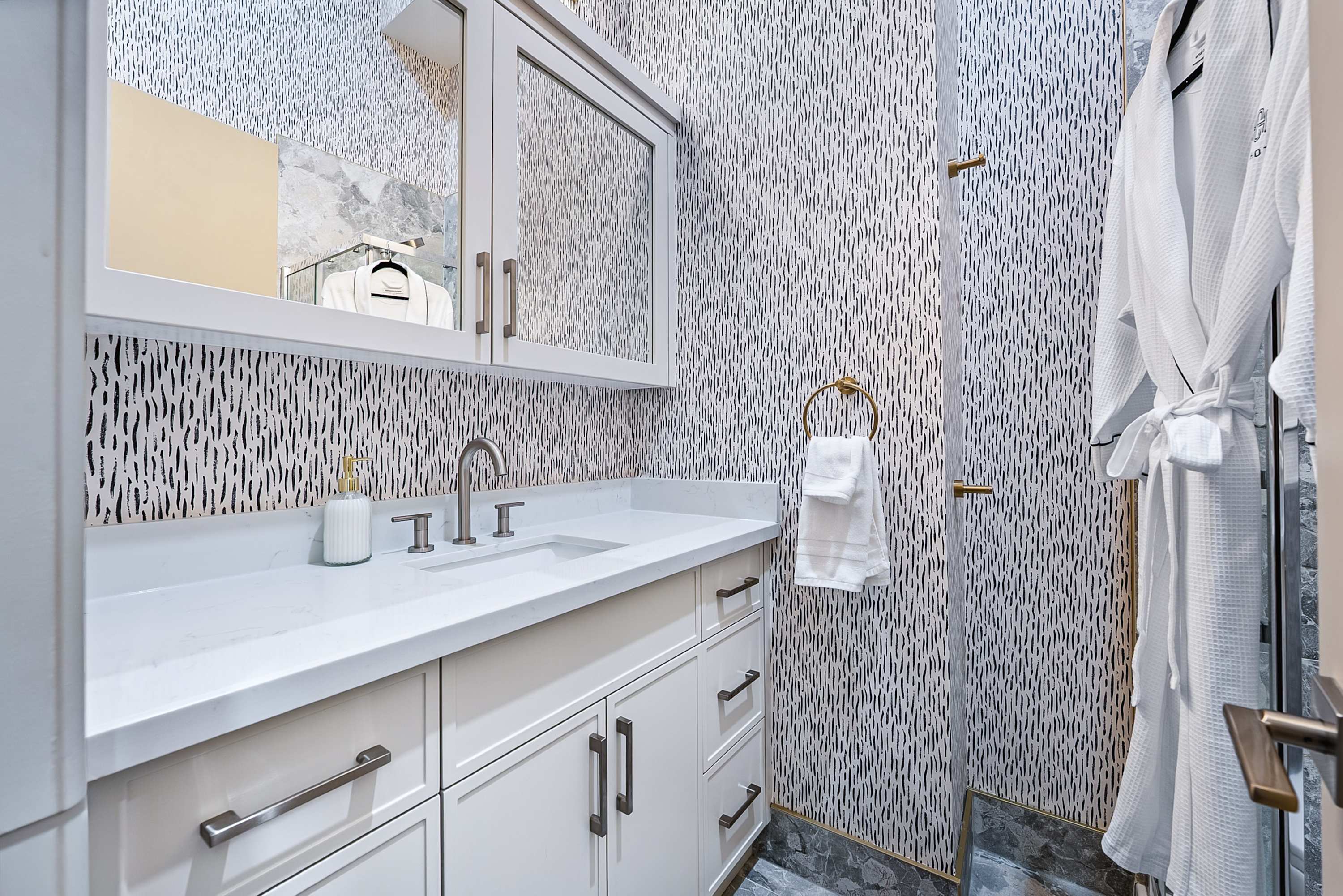 A modern bathroom featuring a white vanity with a rectangular sink, brushed nickel faucet, and mirrored medicine cabinet, set against a black and white patterned wallpaper.
