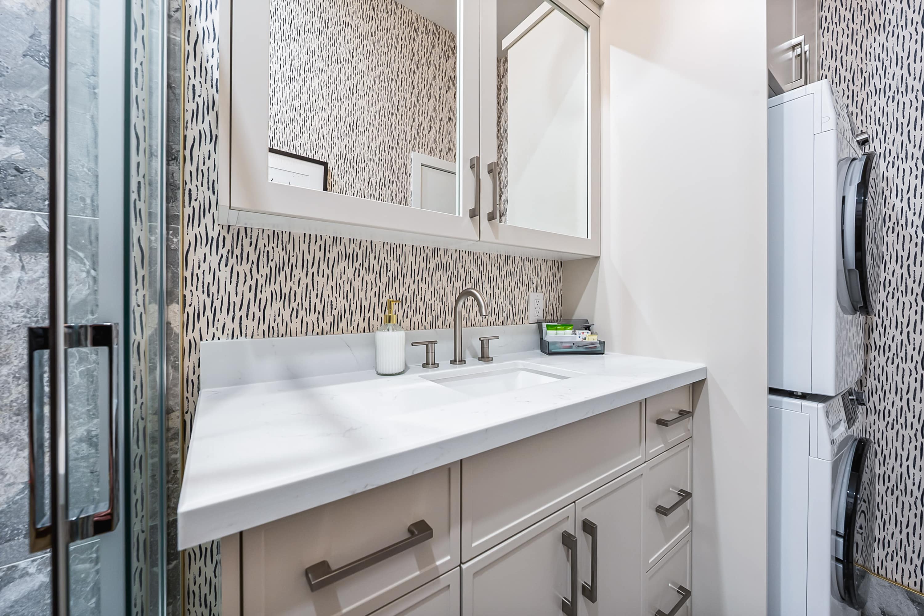A wider view of the bathroom vanity area, revealing a stacked washer and dryer unit next to the white vanity, which has a single sink and mirrored medicine cabinets above.