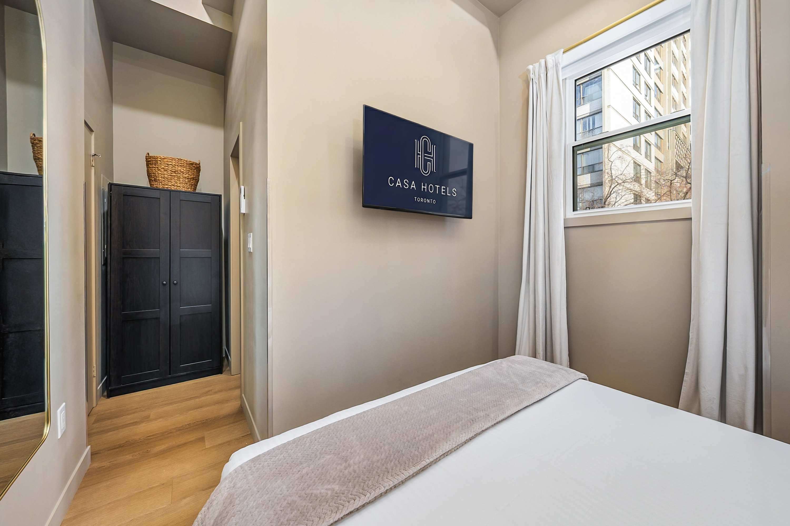 A different angle of the bedroom showing a wall-mounted TV displaying "CASA HOTELS TORONTO" above the foot of the bed, with a dark wardrobe visible in the background through an open doorway.