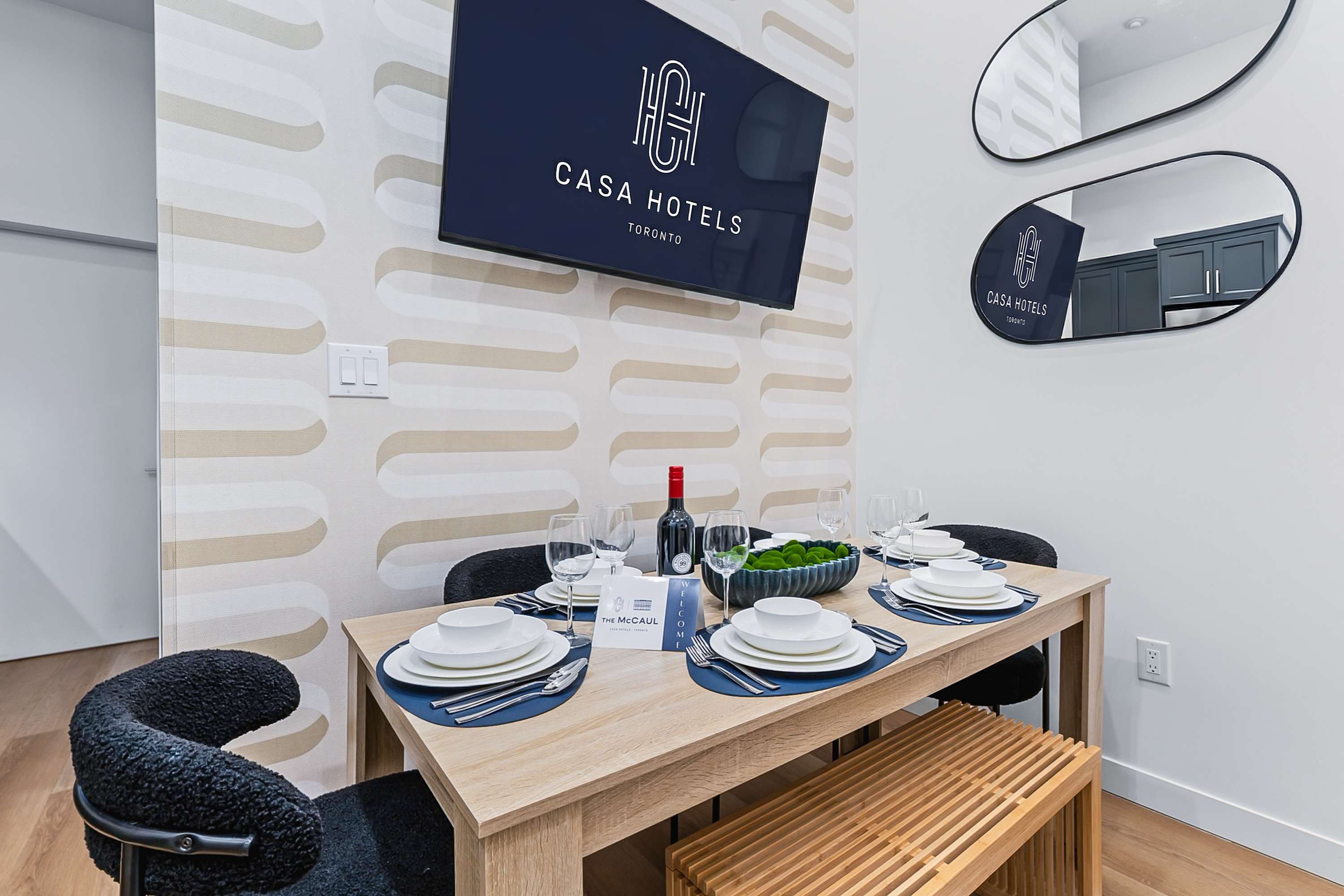 A neatly set dining table made of light wood, prepared with white dinnerware, blue placemats, and wine glasses, positioned against a wall with a distinctive wavy pattern wallpaper.