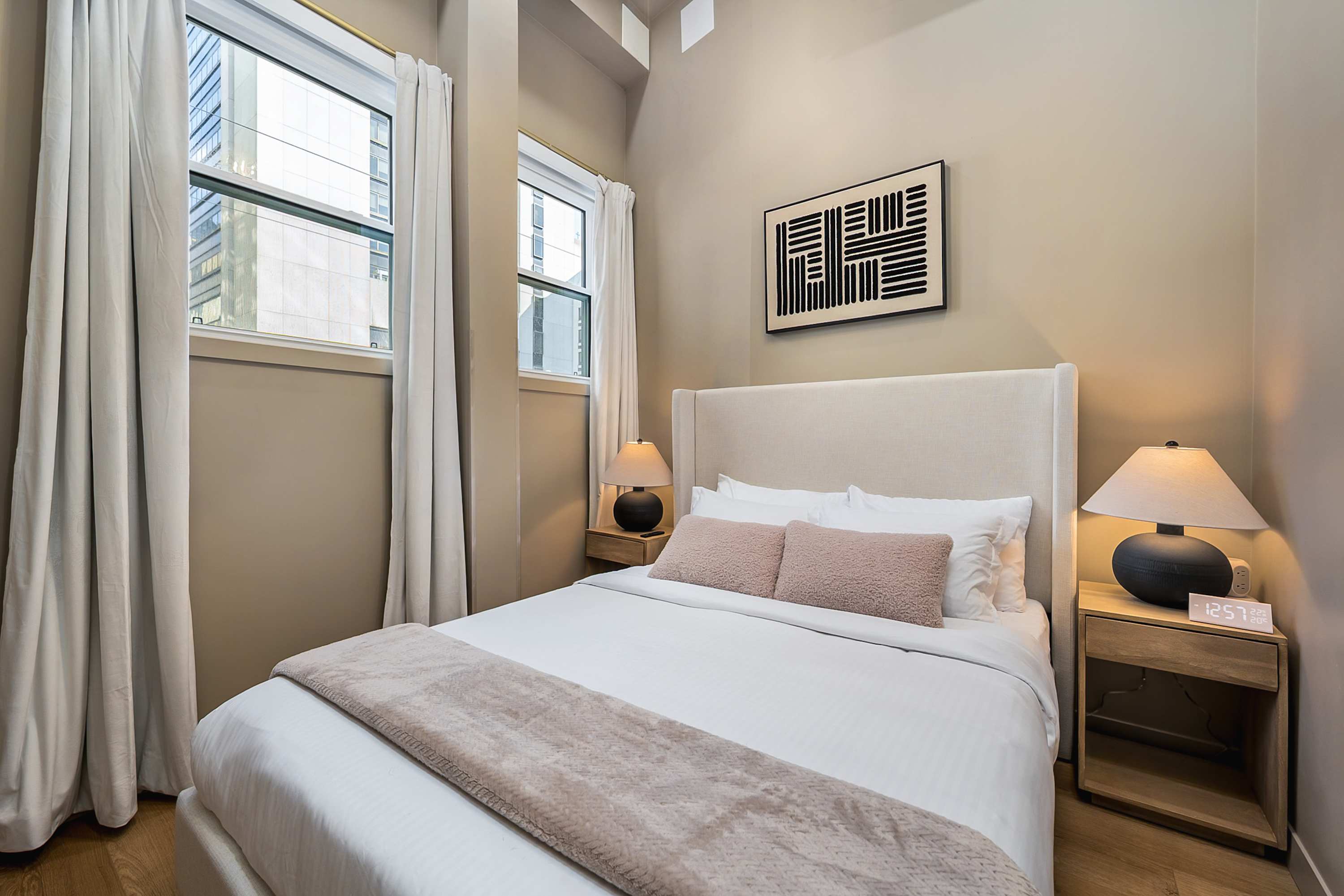 A cozy bedroom featuring a queen-sized bed with a tall upholstered headboard, white bedding, and pink accent pillows, flanked by two wooden nightstands with lamps, and two large windows with white curtains.