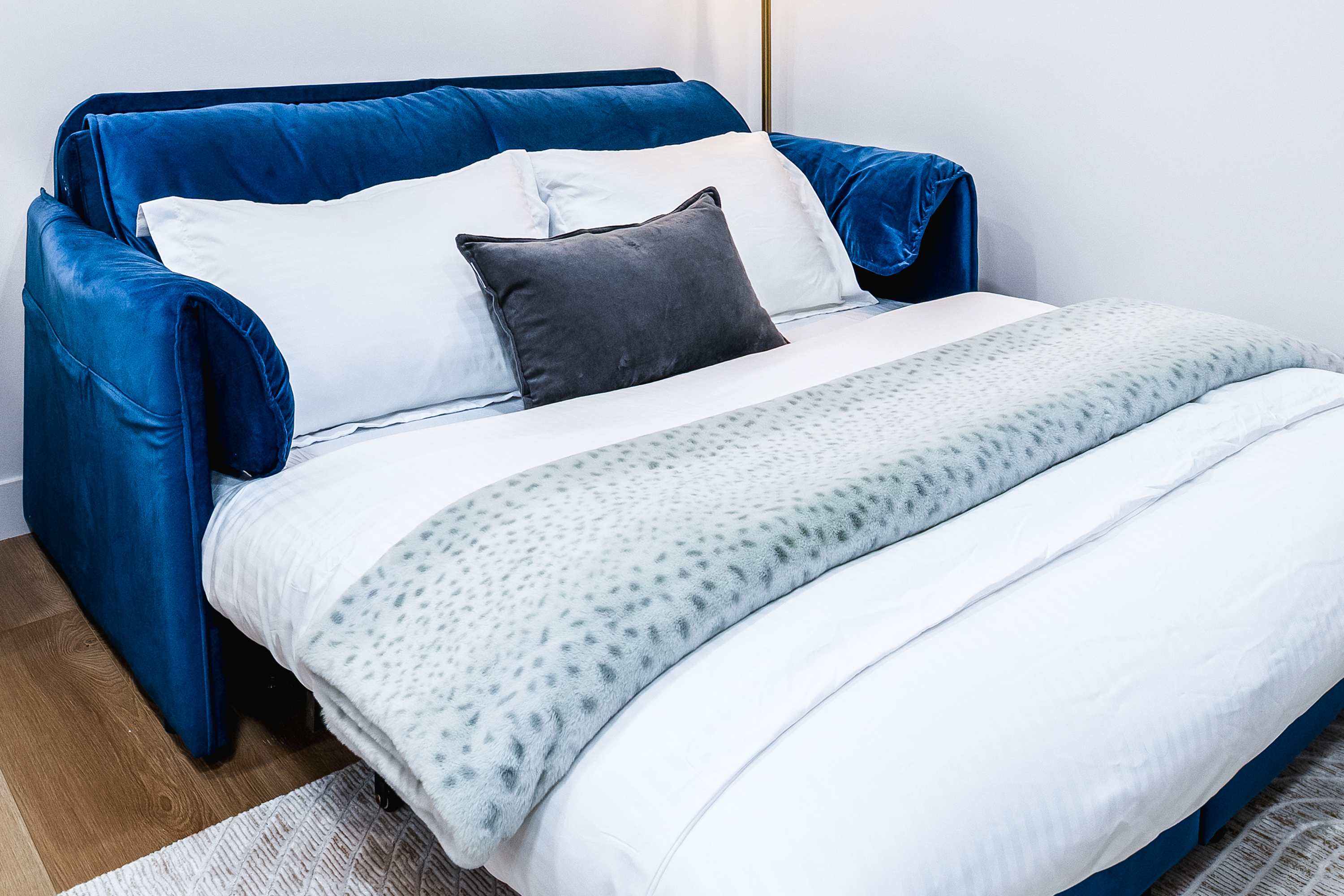 A vibrant blue velvet sofa is transformed into a pull-out bed, dressed with white linens and a soft gray patterned throw blanket. The sofa bed is positioned on light hardwood floors, suggesting a versatile sleeping arrangement in the living area.