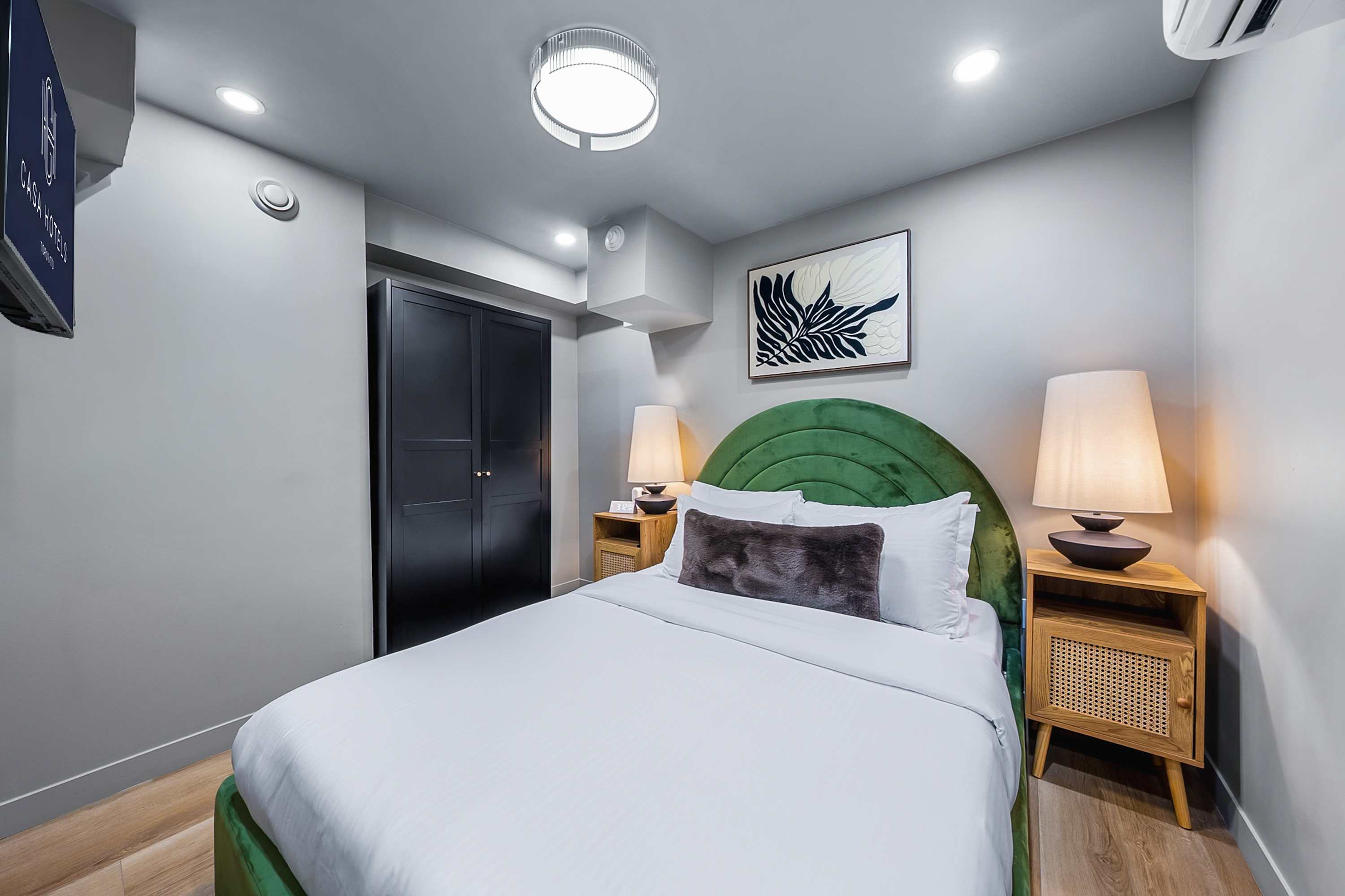 A cozy bedroom features a queen-sized bed with a green velvet headboard and white bedding, flanked by two wooden nightstands with woven cane fronts and modern lamps. A dark wardrobe stands against the light gray wall, beneath a contemporary abstract artwork.