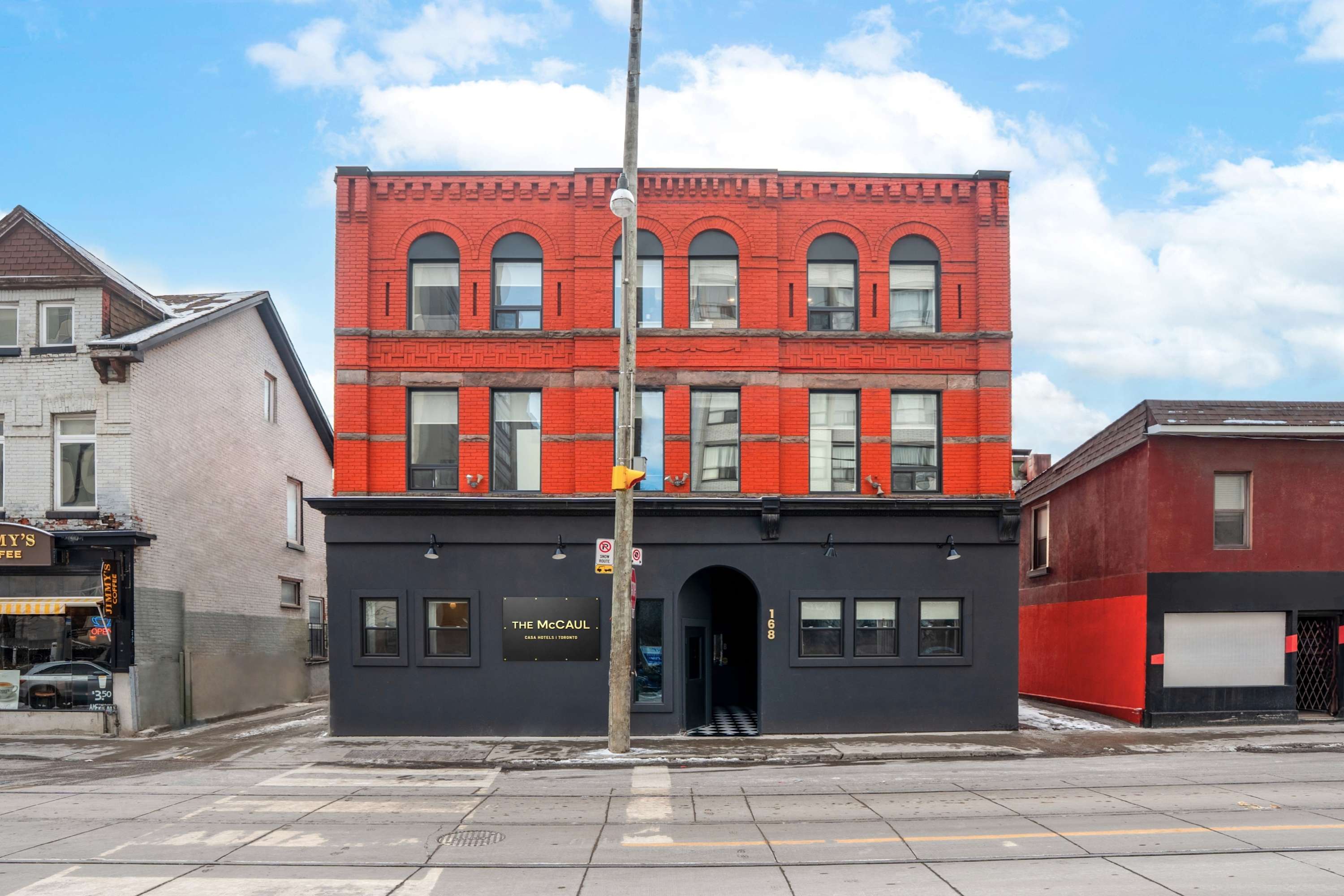 A full frontal view of "THE McCAUL" hotel, a striking red brick building with arched windows on the upper floors and a dark gray ground level. The building stands prominently on a city street with a clear blue sky overhead, showcasing its distinct architectural style.