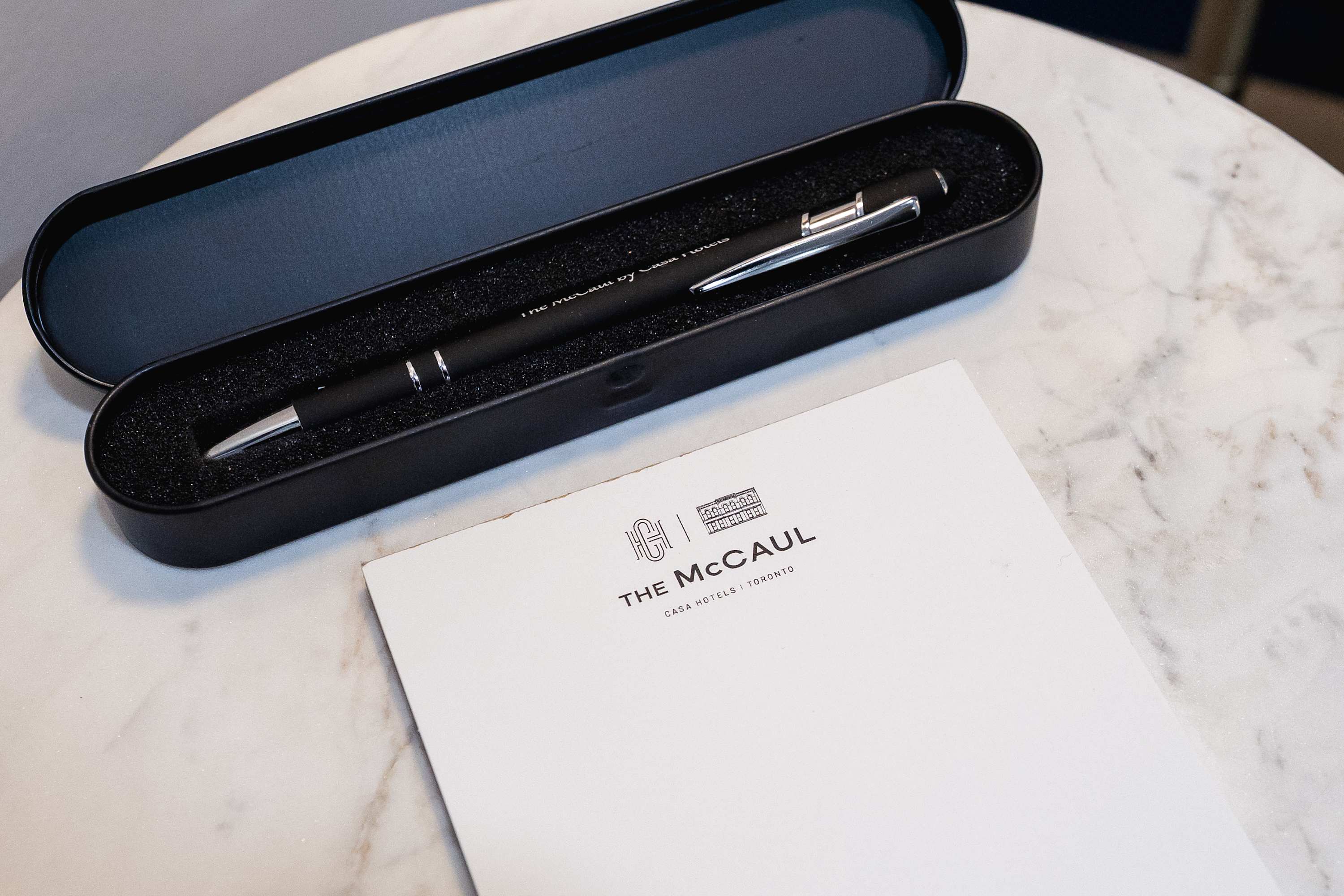 A close-up of a black pen engraved with "The McCaul by Casa Hotels" resting in its case, alongside a notepad featuring "THE McCAUL CASA HOTELS | TORONTO" logo. These items are placed on a light-colored surface, likely a table.