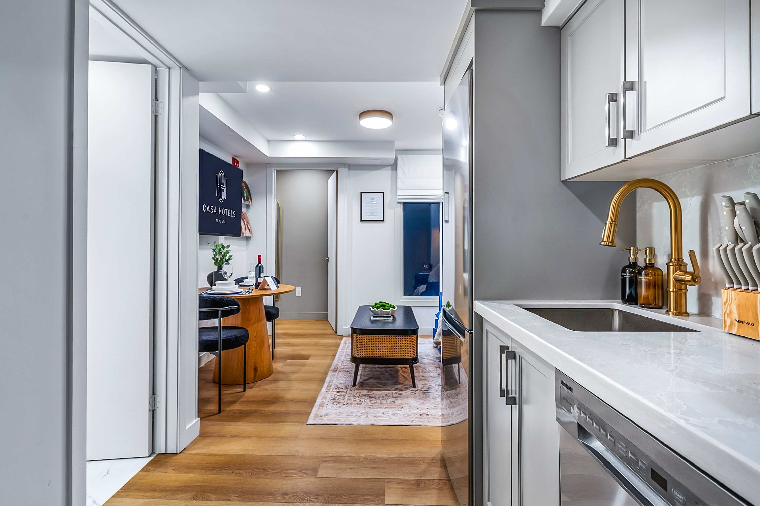 A view from the kitchen into the open-concept living and dining area, featuring warm hardwood floors and light gray walls. A round wooden dining table with black chairs is visible, along with a black coffee table and a blue sofa in the distance.