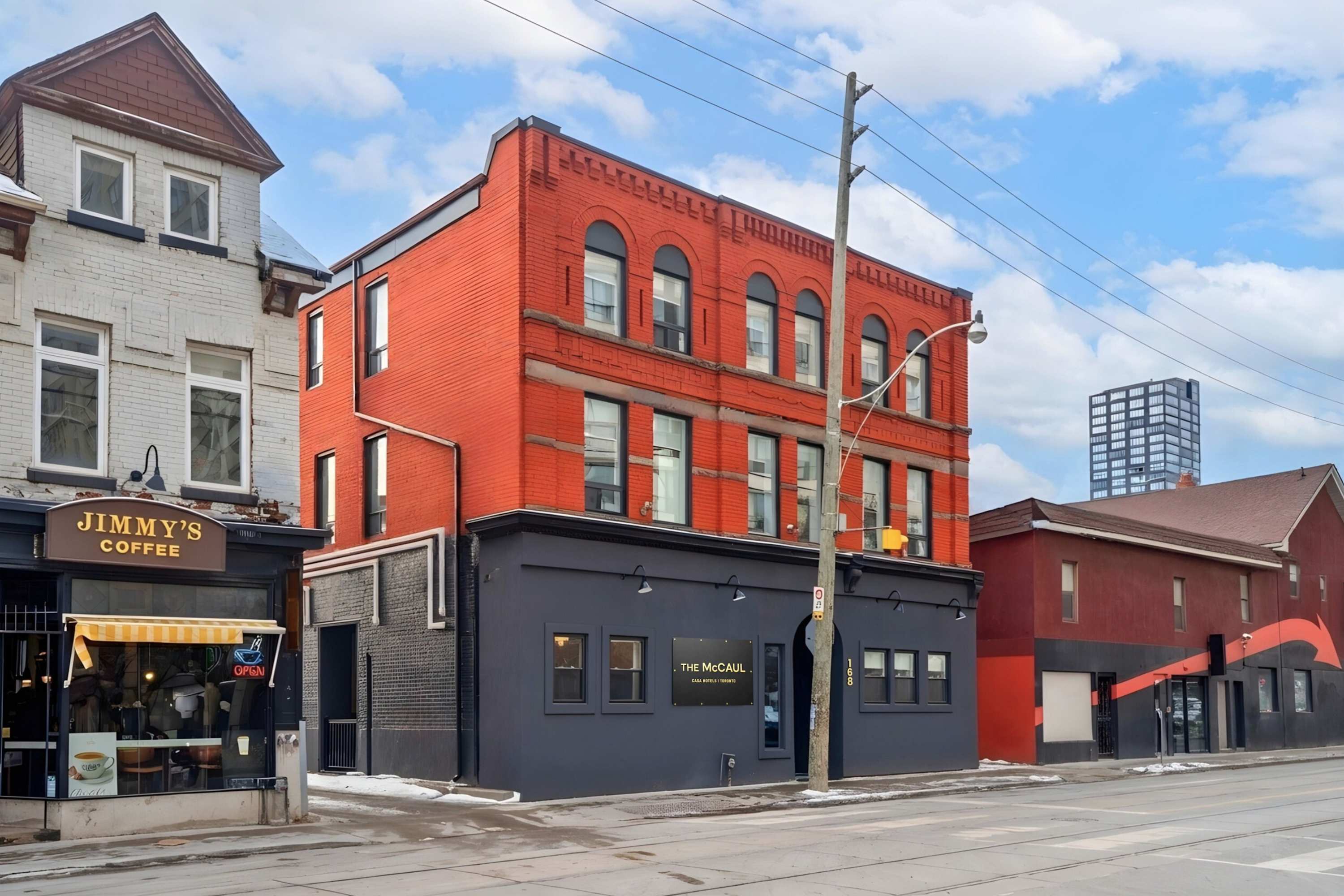 A vibrant red brick building with dark grey lower levels, housing 'The McCaul' hotel and 'Jimmy's Coffee' next door, is seen from a street view under a partly cloudy sky. A prominent utility pole stands in front of the building.