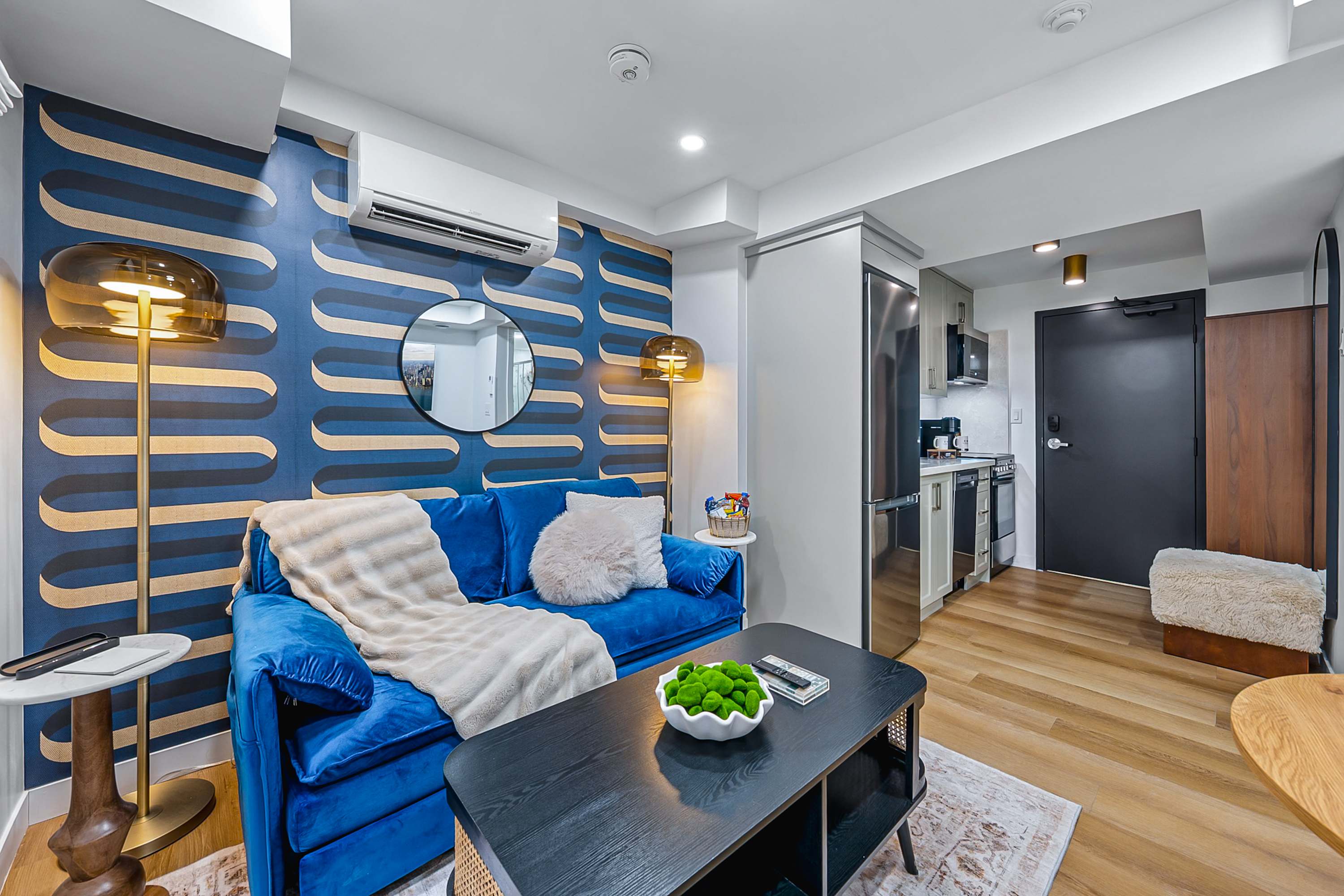 Vibrant living room featuring a royal blue velvet sofa adorned with throw pillows and a blanket, a sleek black coffee table with decorative moss balls, and a striking blue and gold patterned accent wall. Light wood flooring extends into the adjacent kitchen area.