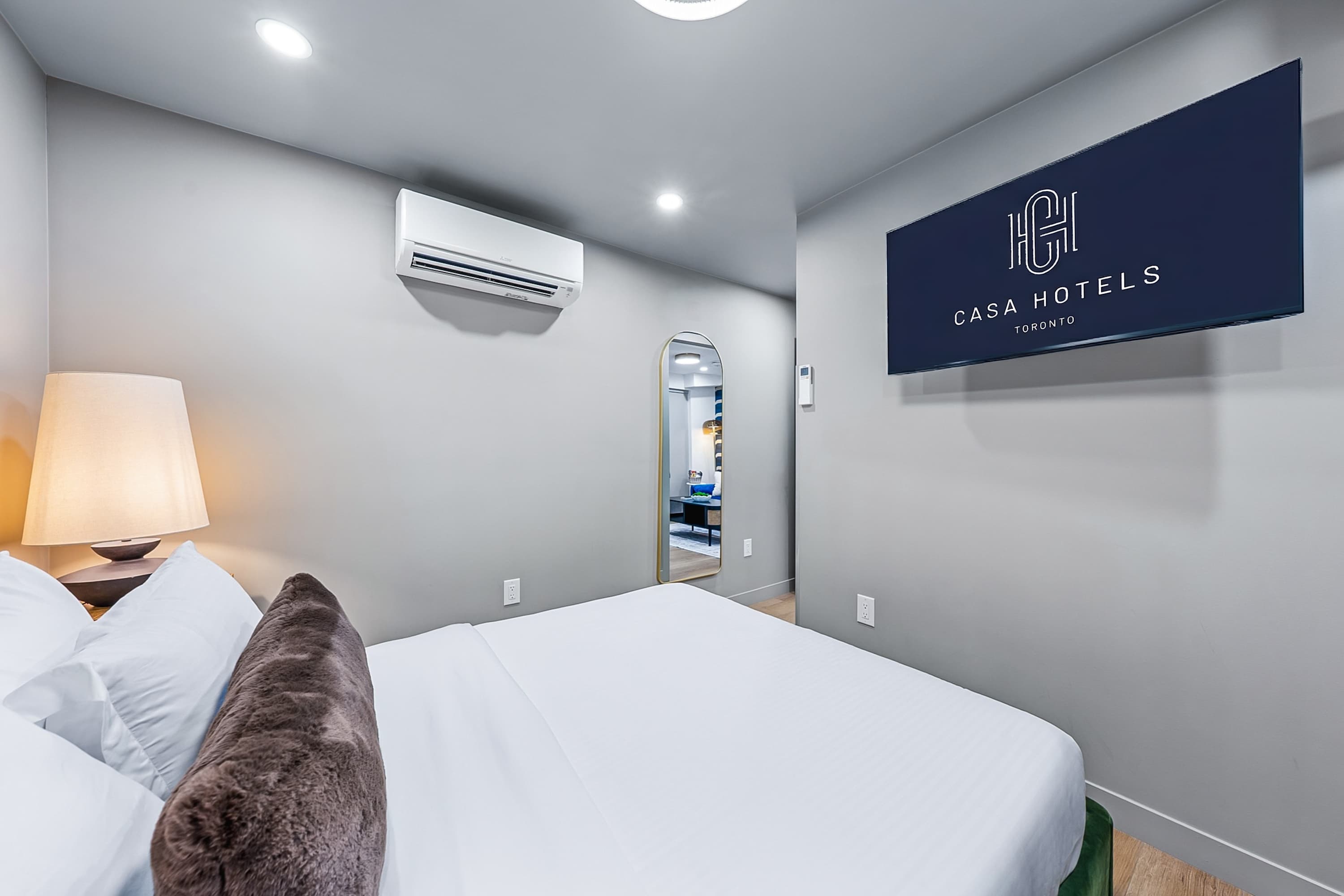 This bedroom showcases a large wall-mounted TV displaying "CASA HOTELS Toronto" above a queen bed with white linens and a green velvet headboard. An air conditioning unit is mounted high on the light gray wall, and a full-length arched mirror reflects the adjacent living space.