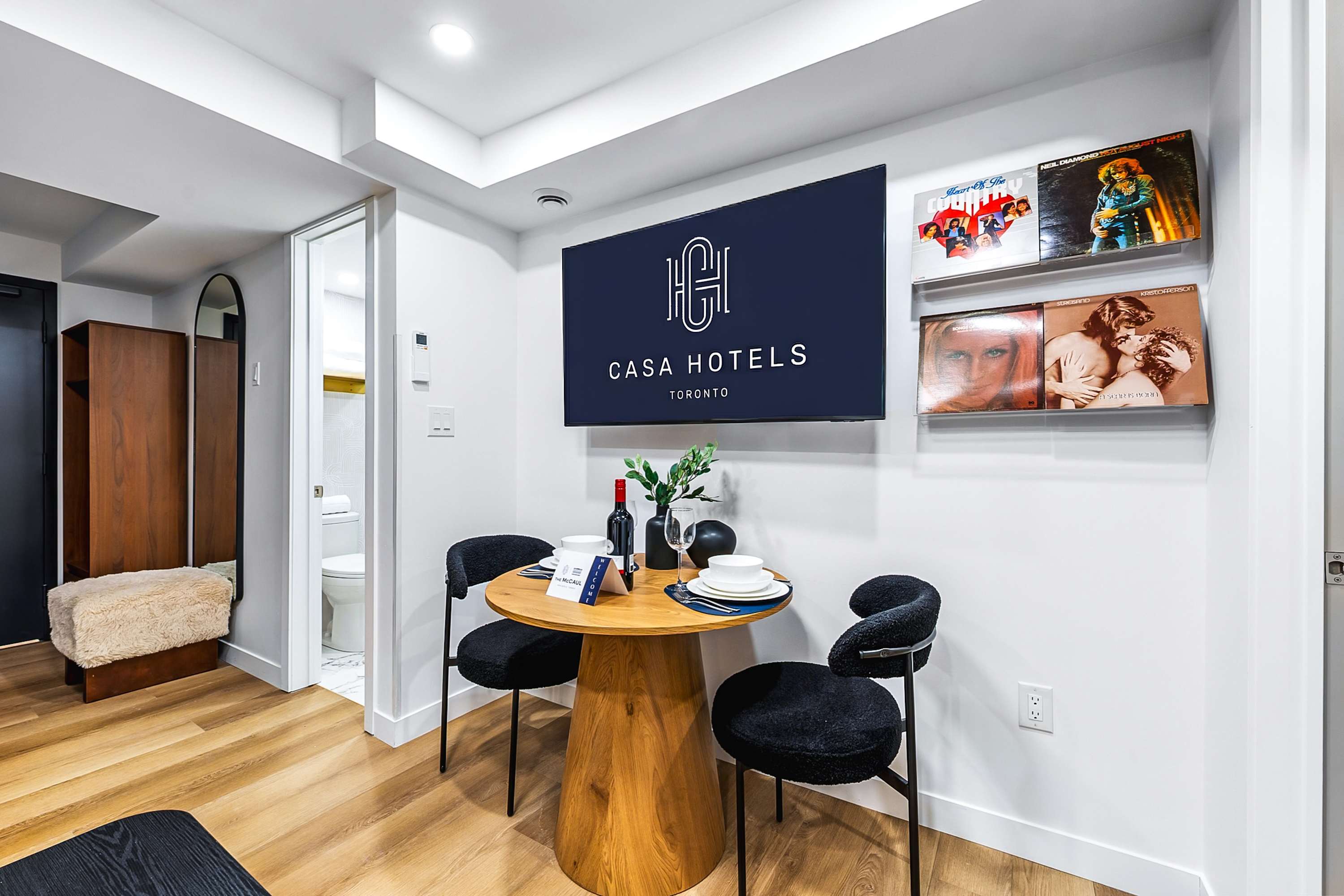 Dining area with a round wooden table and two black textured chairs, positioned beneath a wall-mounted TV displaying 'CASA HOTELS TORONTO'. Decorative vinyl records are stylishly displayed on clear shelves to the right of the TV.