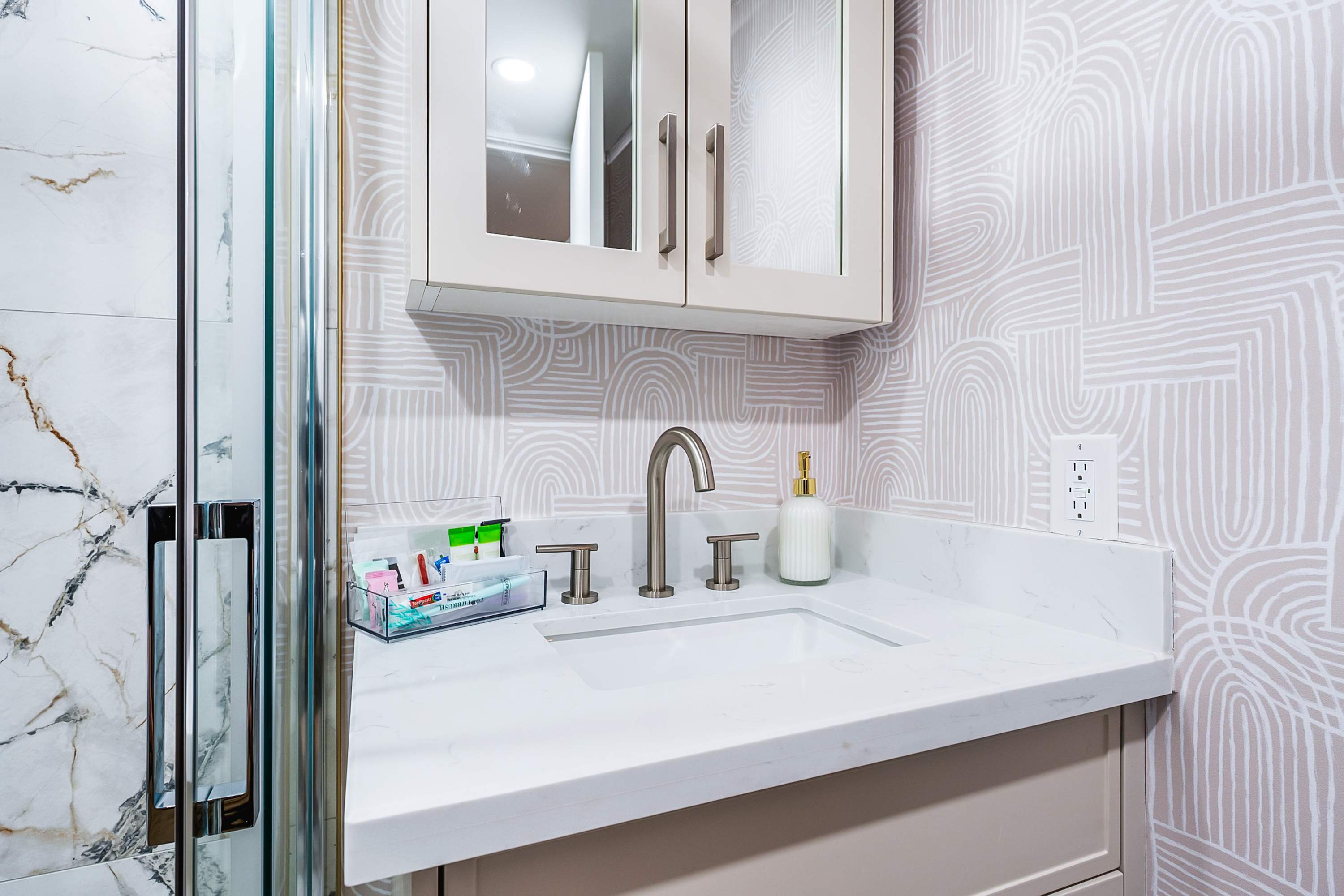 A modern bathroom features a white vanity with a rectangular sink and brushed nickel faucet, set against a light-patterned wallpaper. A mirrored medicine cabinet hangs above, and a glass shower enclosure with marble-patterned tiles is visible to the left.