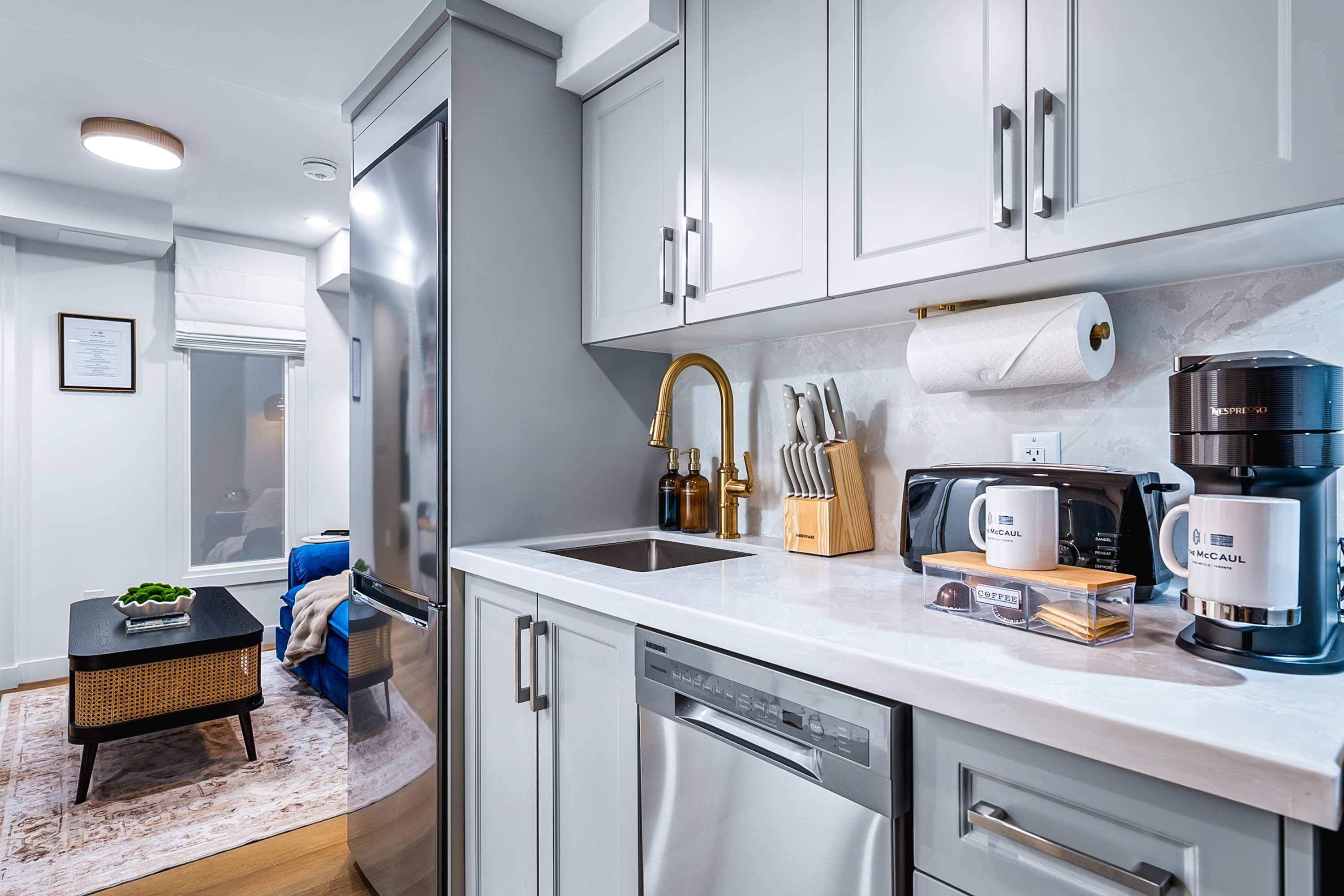 A well-equipped kitchen showcases light gray shaker-style cabinets, a white marble-patterned countertop, and a gold gooseneck faucet over a stainless steel sink. Appliances include a coffee maker, toaster, and a knife block, with hardwood flooring extending into the adjacent living space.