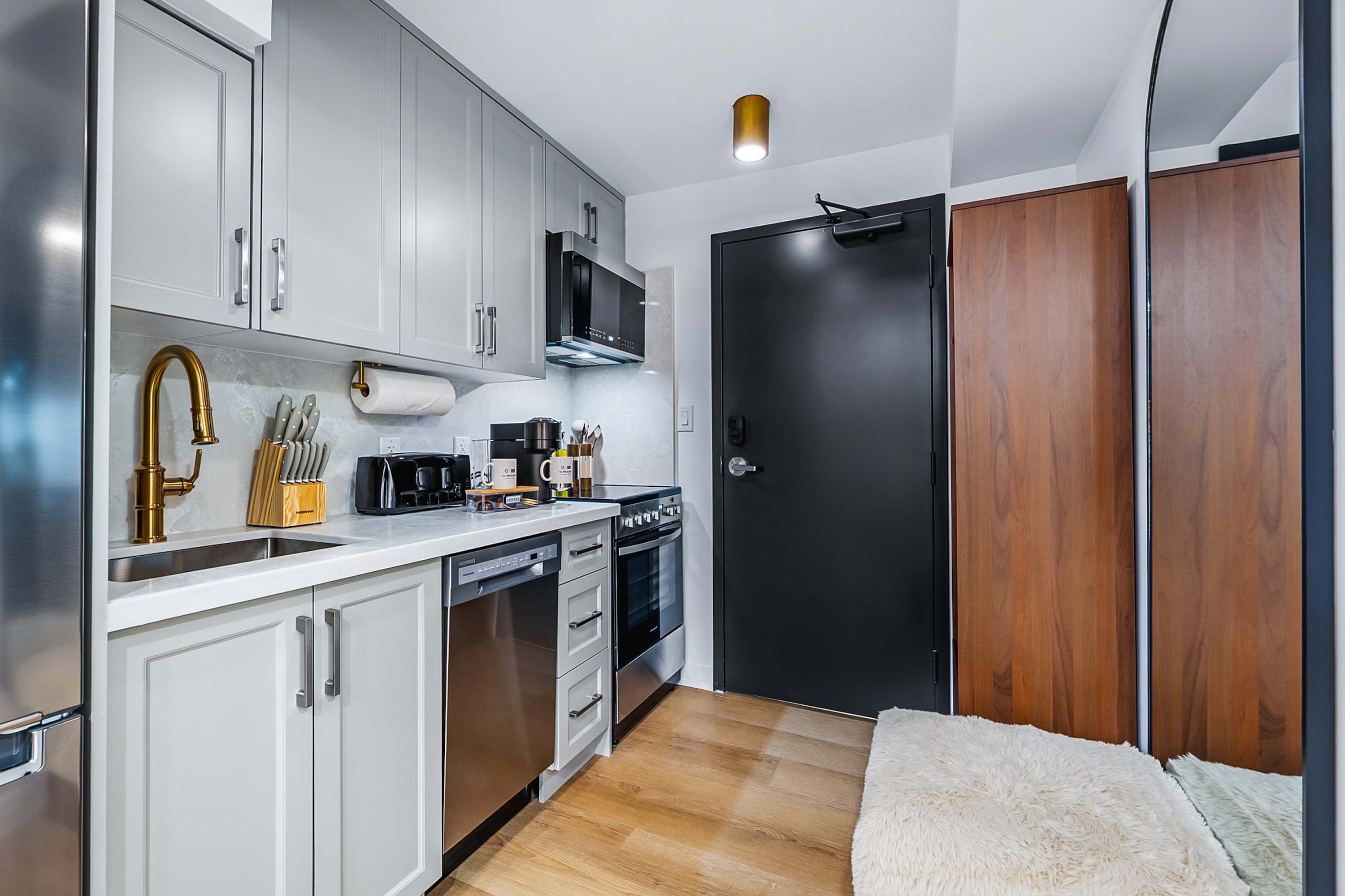 Full view of the compact kitchen featuring light gray shaker cabinets, white countertops, and stainless steel appliances including a refrigerator, stove, and microwave. A gold faucet and various kitchen tools are neatly arranged, with a dark entry door visible on the right.