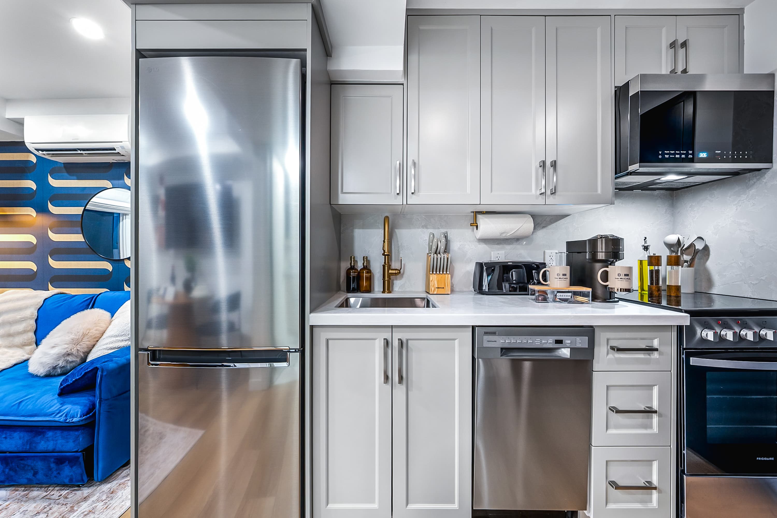A modern, compact kitchen features light gray cabinetry, stainless steel appliances including a refrigerator, dishwasher, and microwave, and a white countertop with a gold-toned faucet. A glimpse of a blue sofa and patterned wall is visible in the adjacent living area.