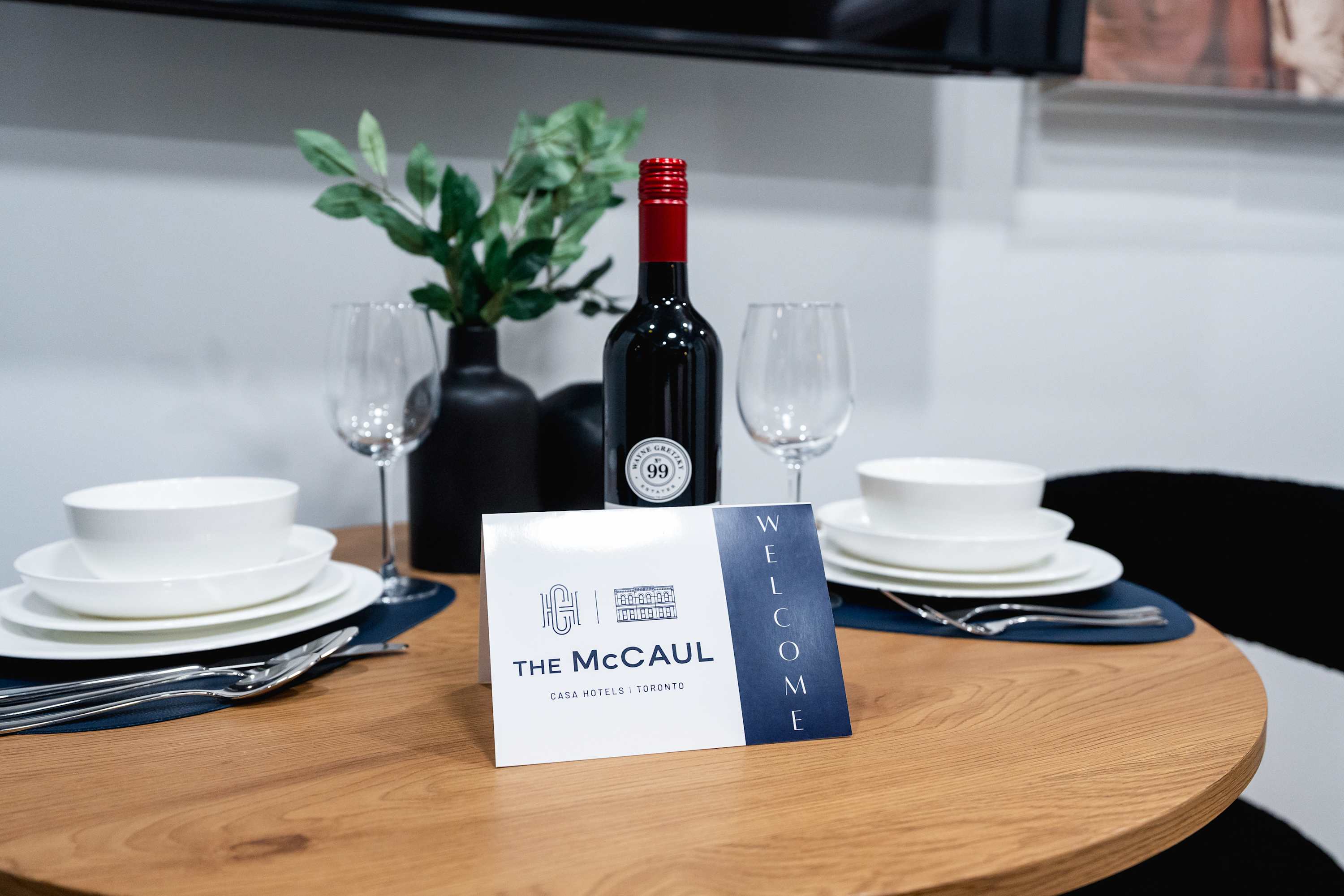 Close-up of the dining table set with white dishes, wine glasses, and a bottle of red wine, alongside a 'WELCOME' card for 'The McCaul | CASA HOTELS TORONTO'. A small green plant in a black vase adds a touch of greenery to the inviting setup.