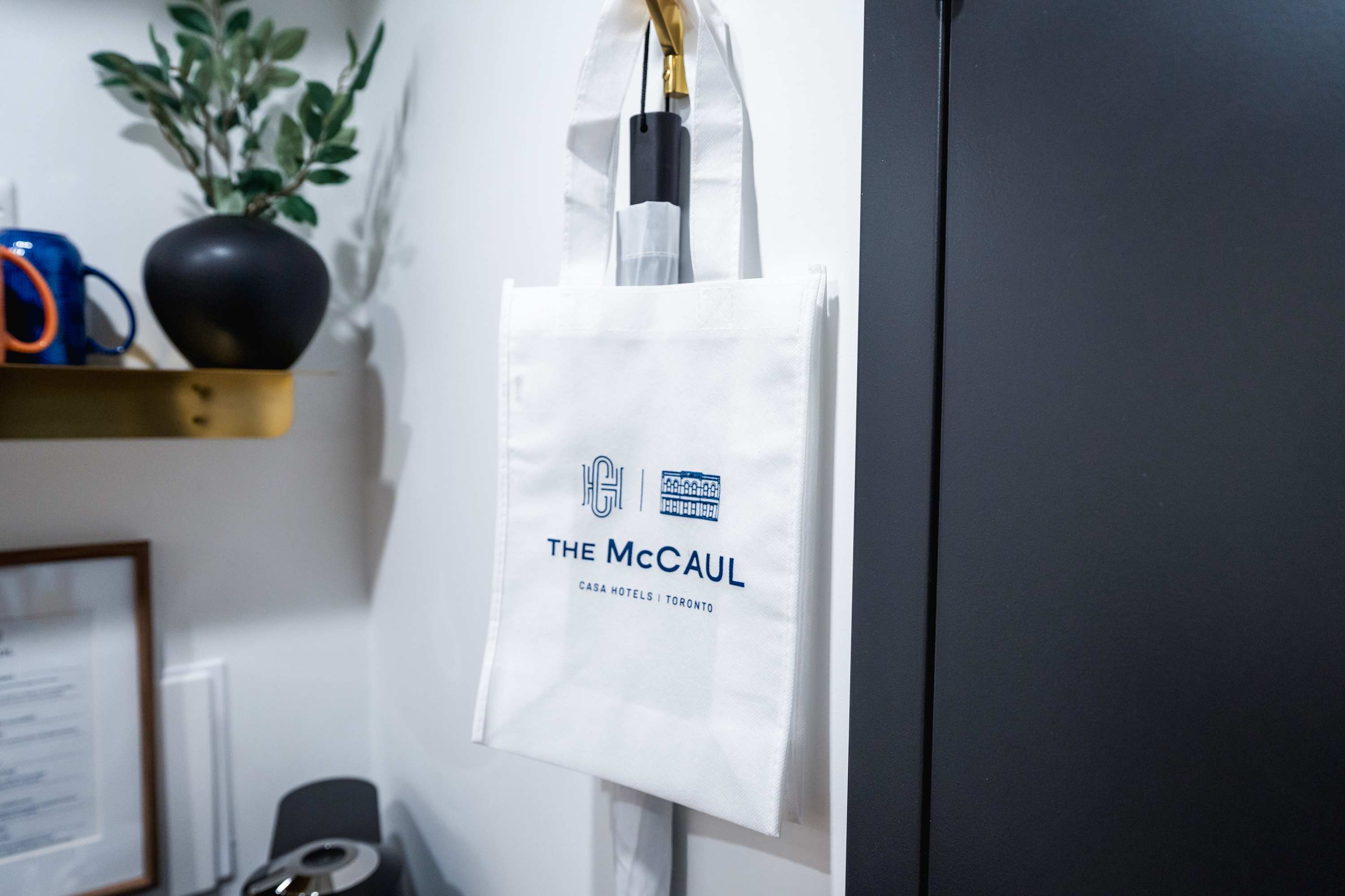 A close-up of a white reusable shopping bag with "THE McCAUL CASA HOTELS | TORONTO" logo, hanging on a hook next to a black door.