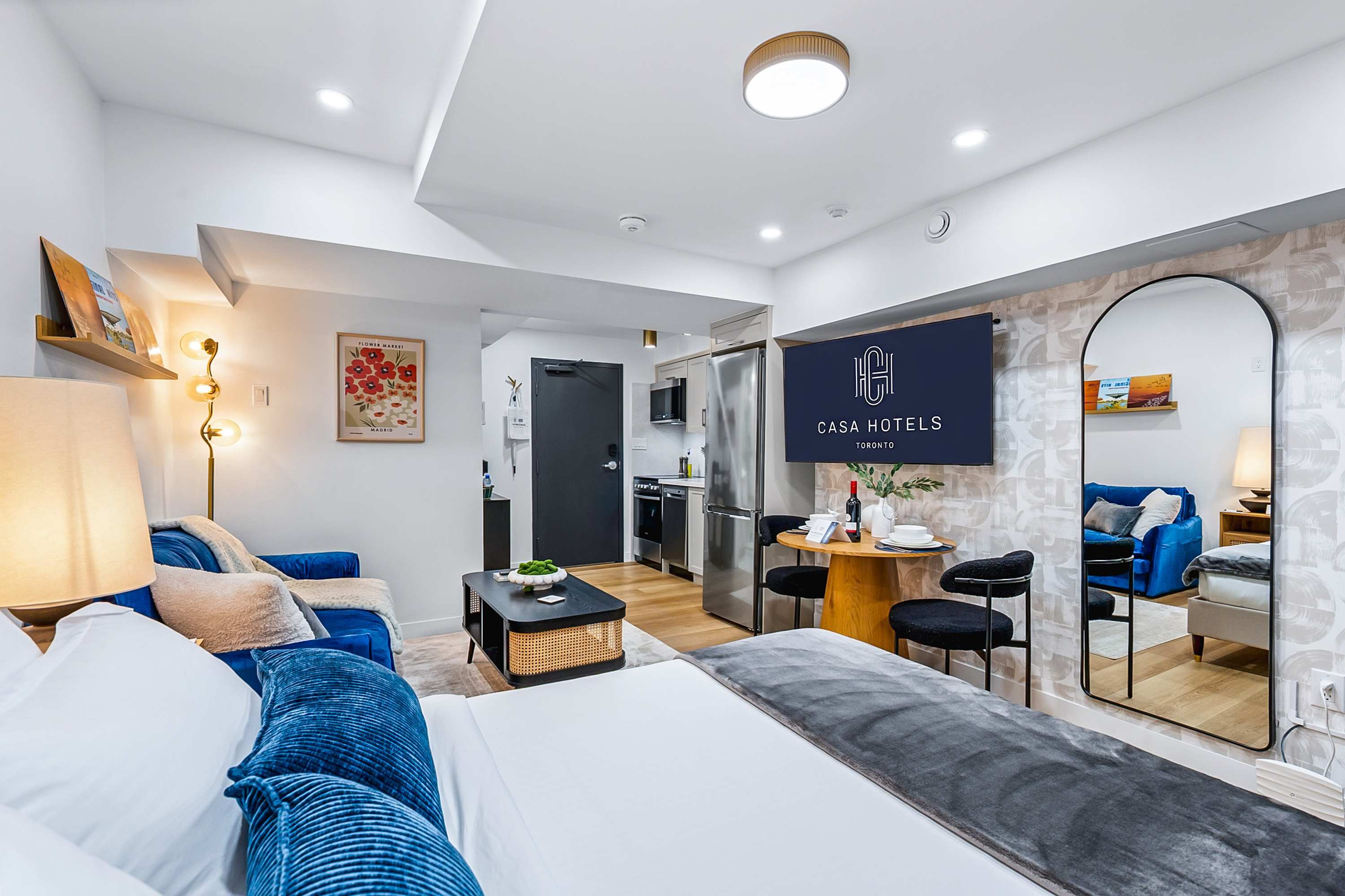 A wide shot of the studio apartment, showcasing the queen bed with blue pillows, a blue velvet sofa, and a black coffee table. A wall-mounted TV with "CASA HOTELS" logo is visible above a small dining table, with a compact kitchen in the background.