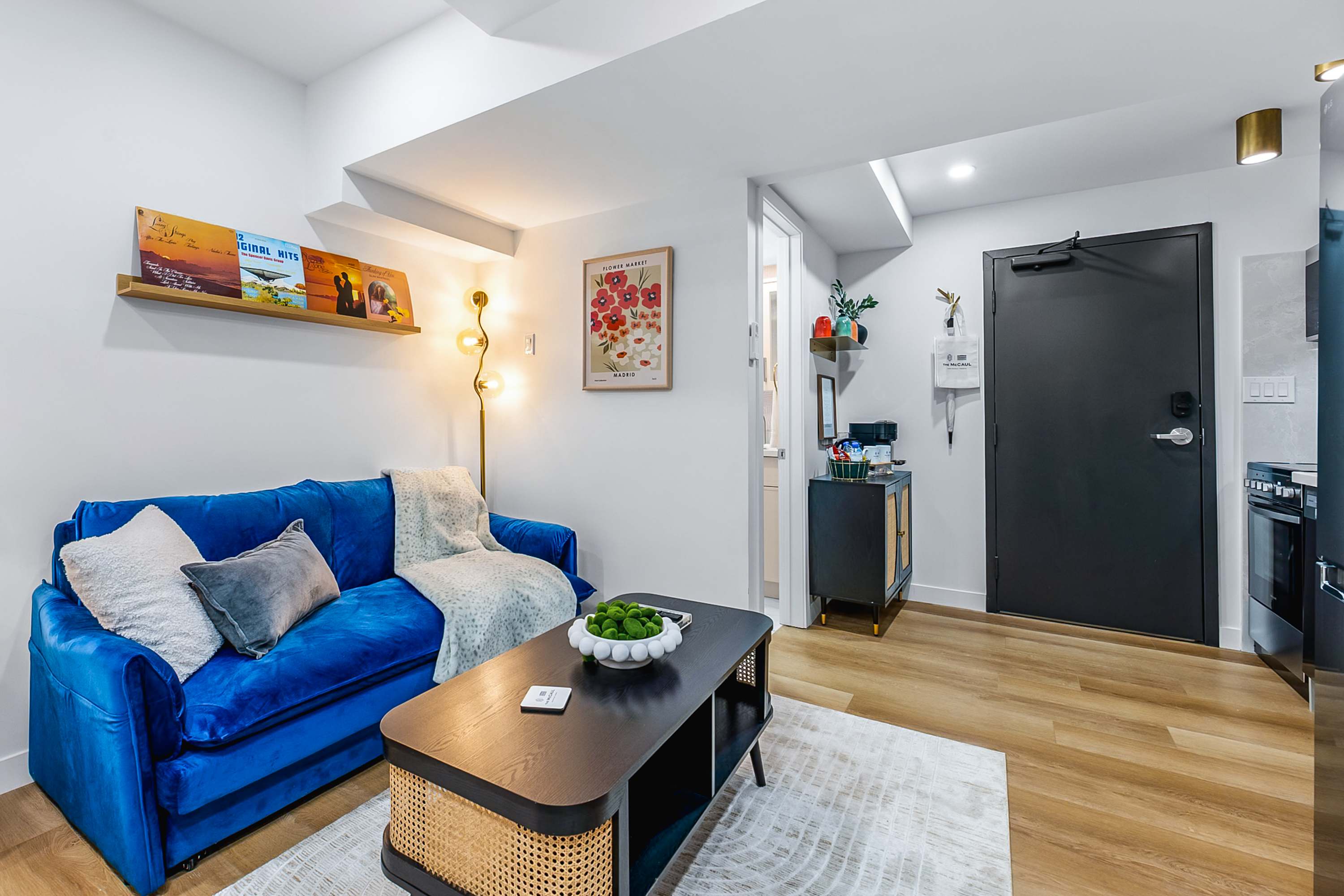 A comfortable living area with a vibrant blue velvet sofa adorned with pillows and a throw, facing a black coffee table with rattan details. A floor lamp provides ambient lighting, and a small bar cart is visible near the entrance.
