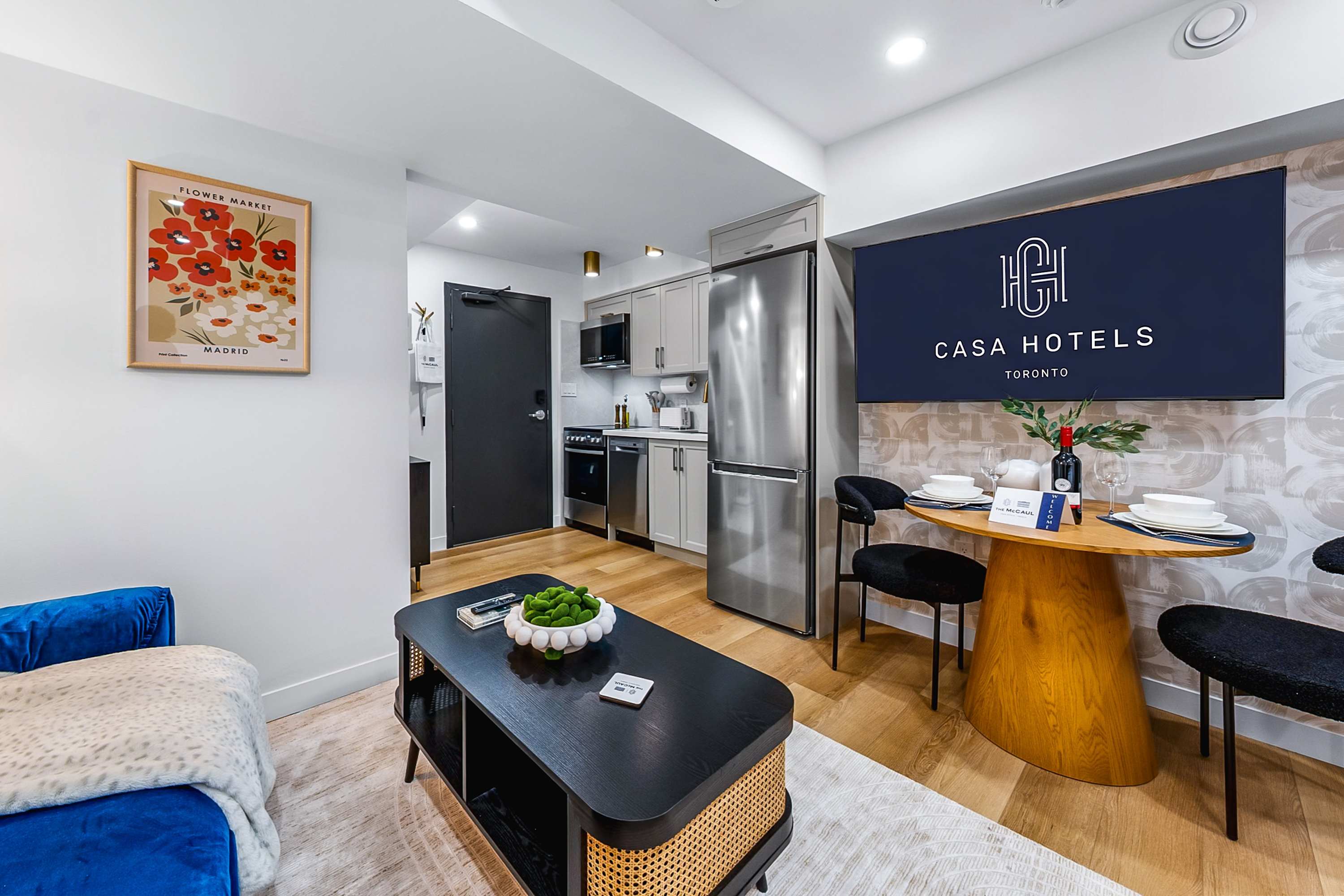 A perspective from the bed, showing the black coffee table, blue velvet sofa, and the dining area with a wall-mounted TV. The compact kitchen with stainless steel appliances is visible in the background.