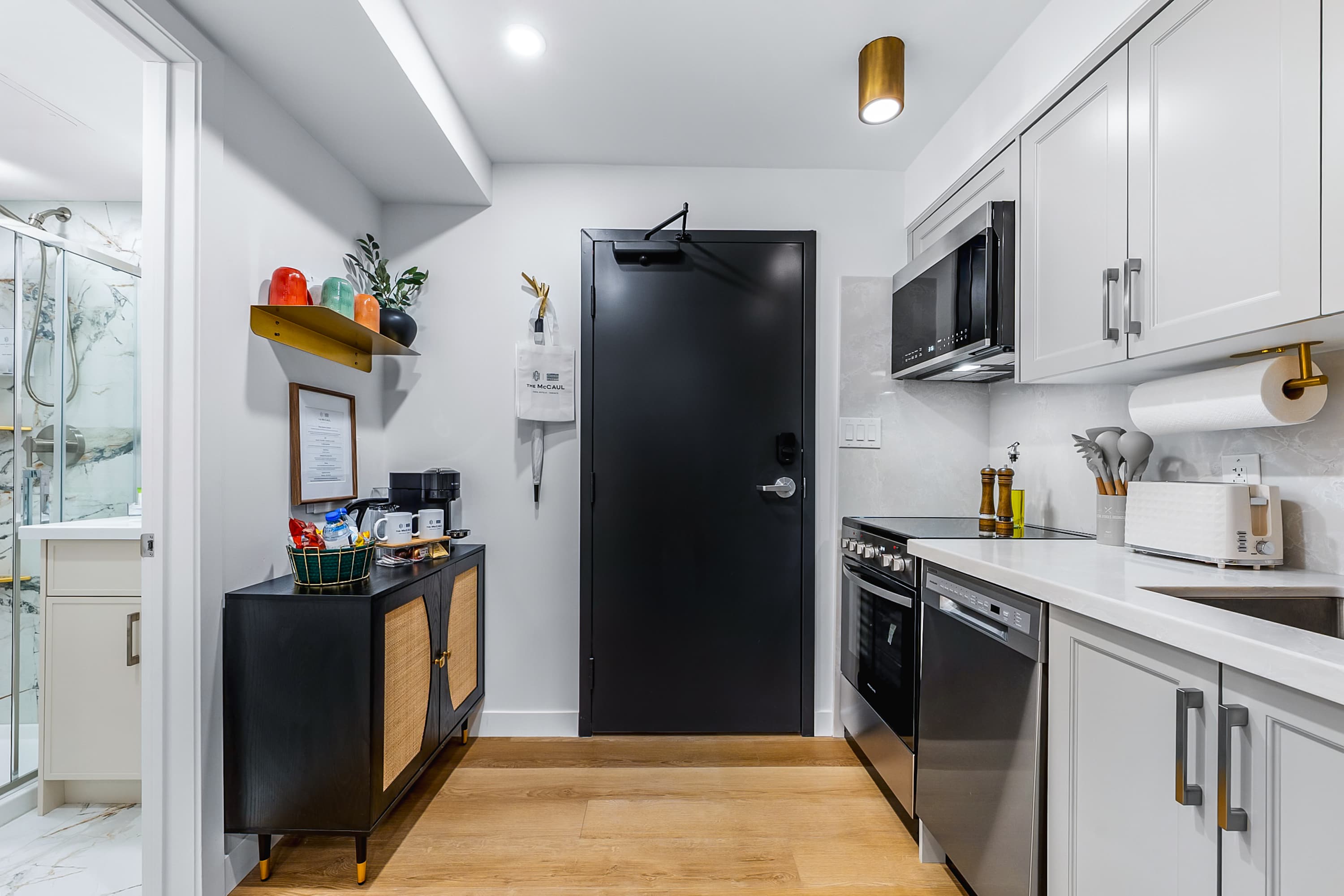 A compact kitchen area with light gray cabinets, white countertops, stainless steel appliances including a stove, microwave, and dishwasher, and a black entry door. A small black cabinet with a coffee maker and snacks is on the left.