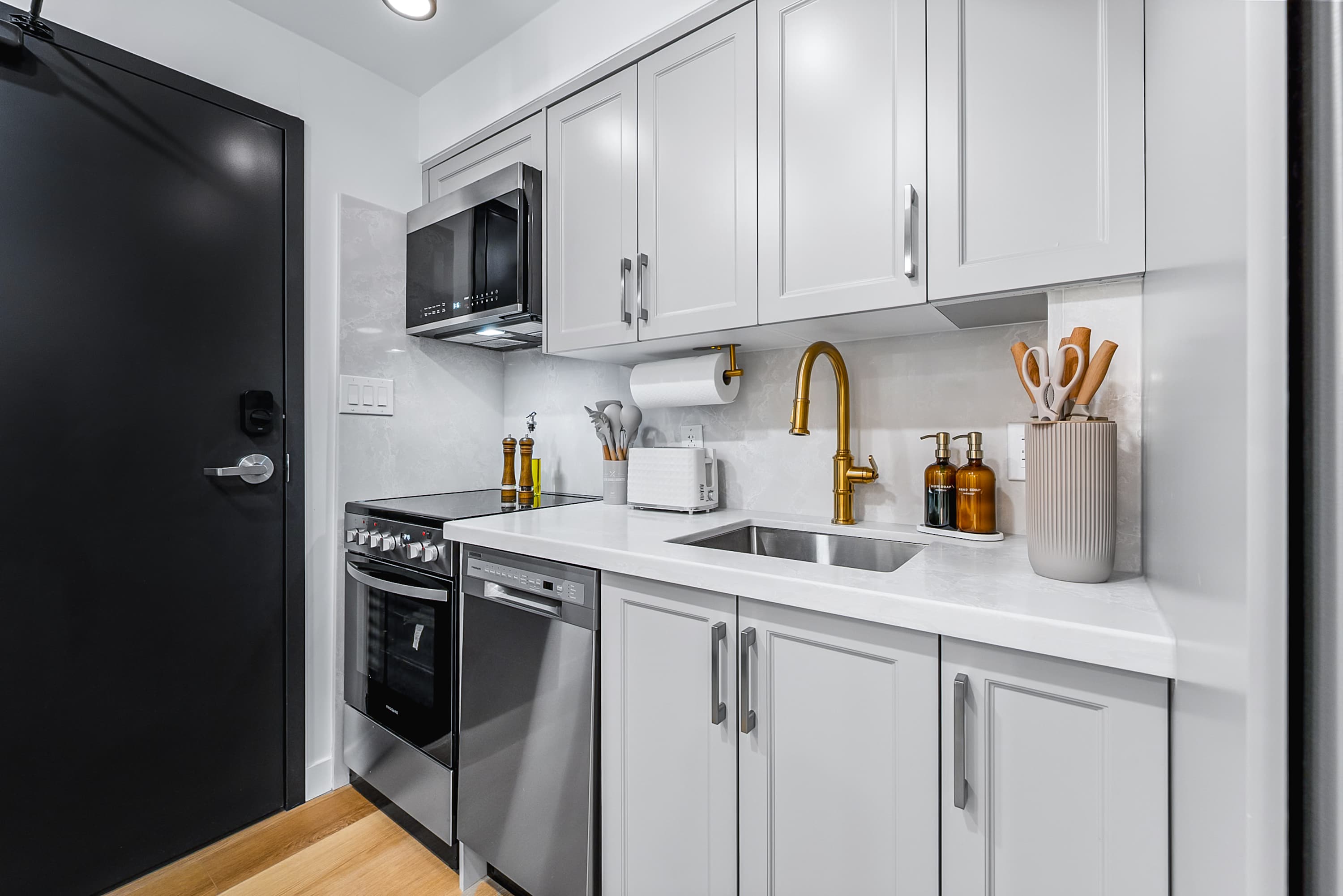 A full view of the modern kitchen featuring light gray shaker-style cabinets, white quartz countertops, stainless steel appliances, and a gold-toned faucet and paper towel holder.
