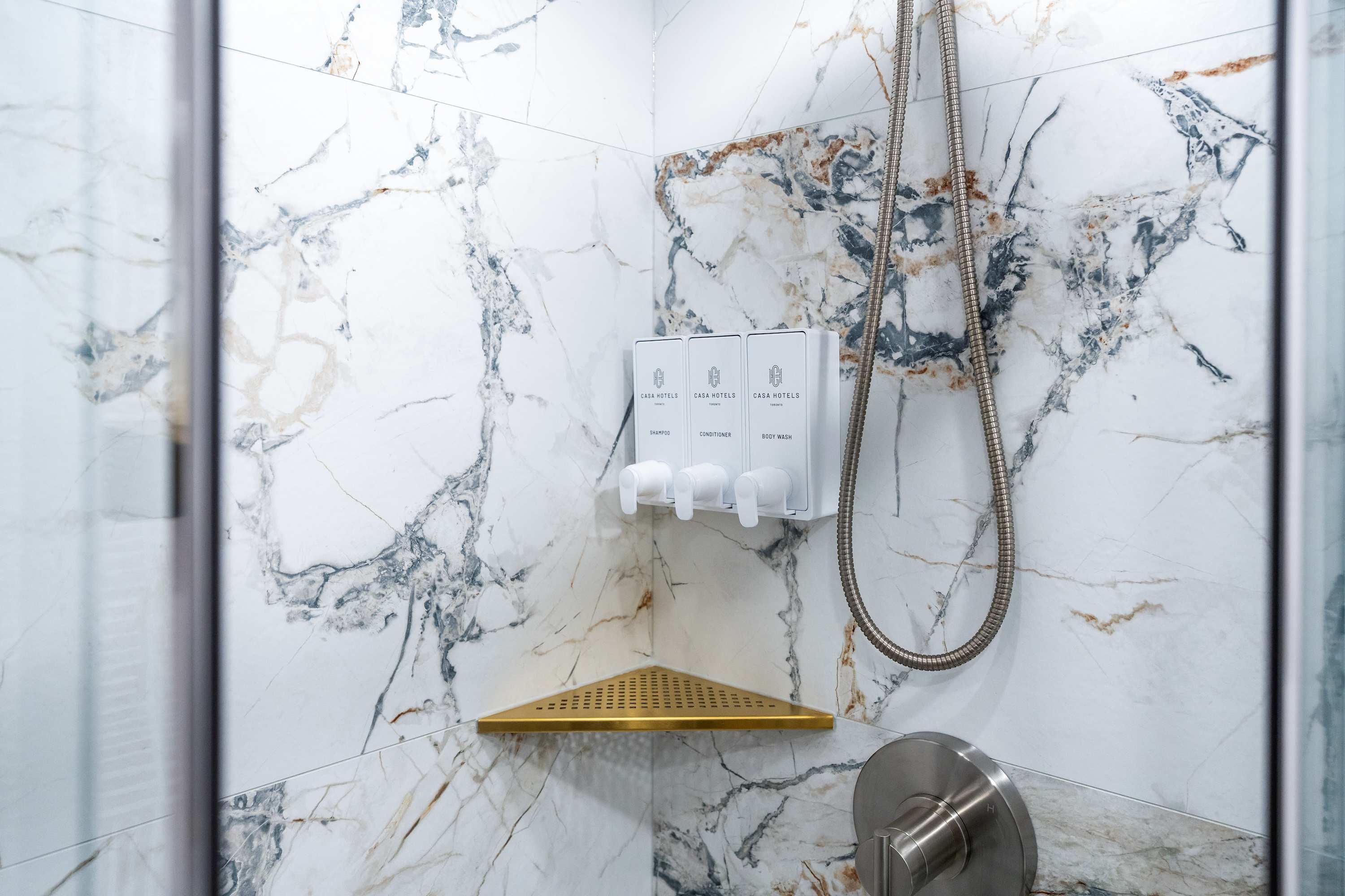 A close-up of a walk-in shower with white and gray marble-patterned tiles, featuring a gold corner shelf and three wall-mounted dispensers for shampoo, conditioner, and body wash with "CASA HOTELS" branding.