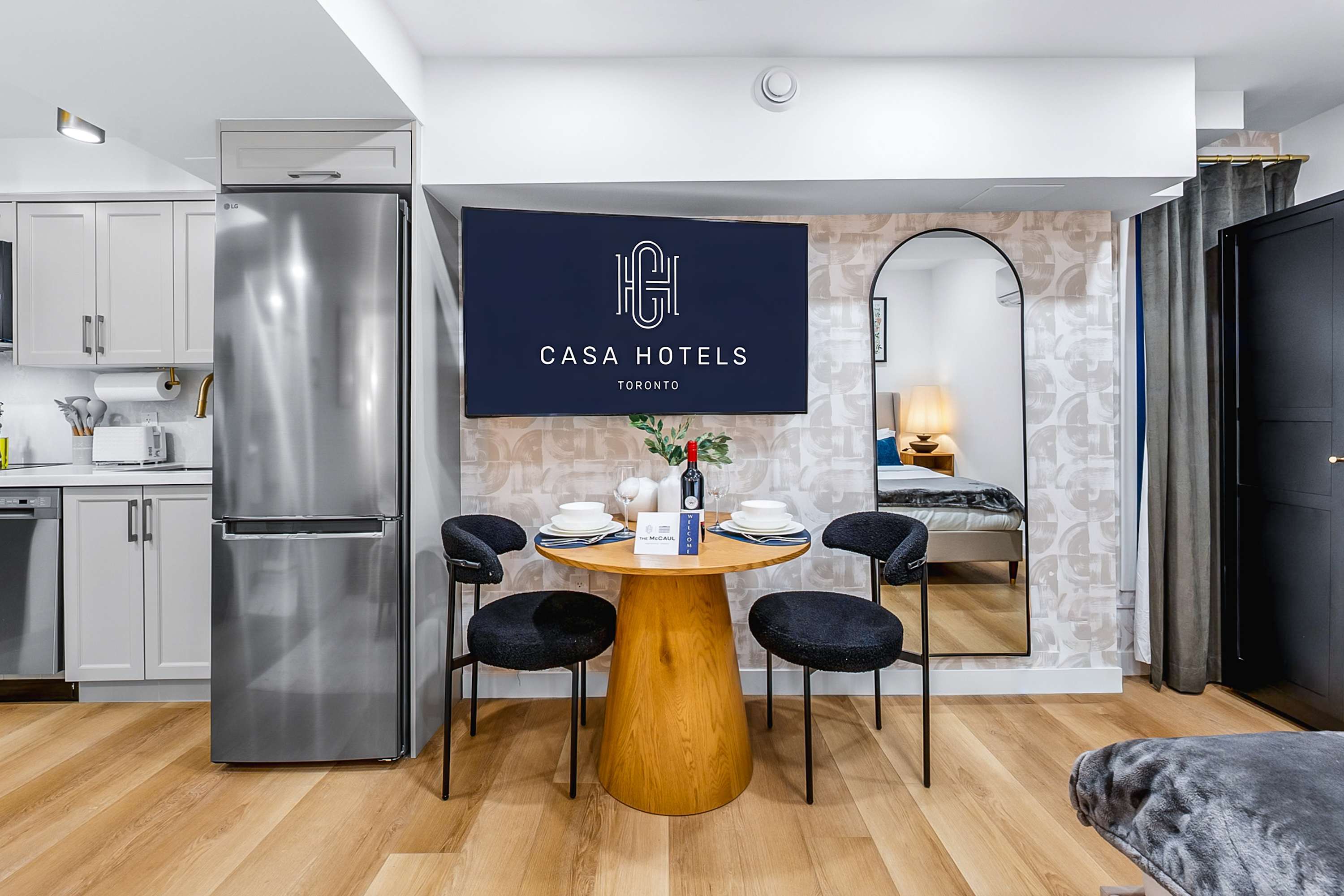 A clear view of the dining area with a round wooden table and two black chairs, positioned in front of a patterned accent wall and a large wall-mounted TV. The modern kitchen with light grey cabinets and stainless steel appliances is to the left.