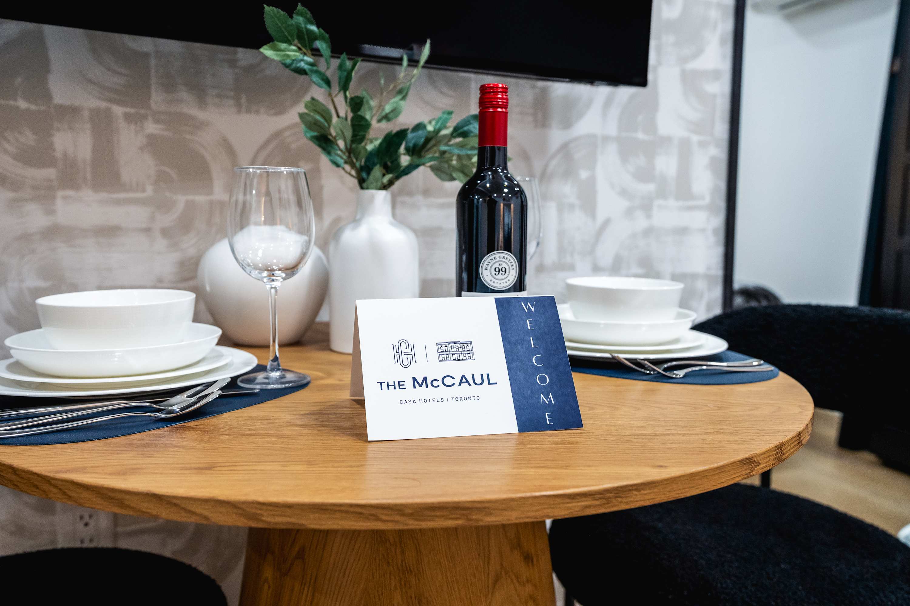 A close-up of a round wooden dining table set for two, featuring white dinnerware, wine glasses, and a bottle of red wine, with a welcome card for "The McCaul" hotel.