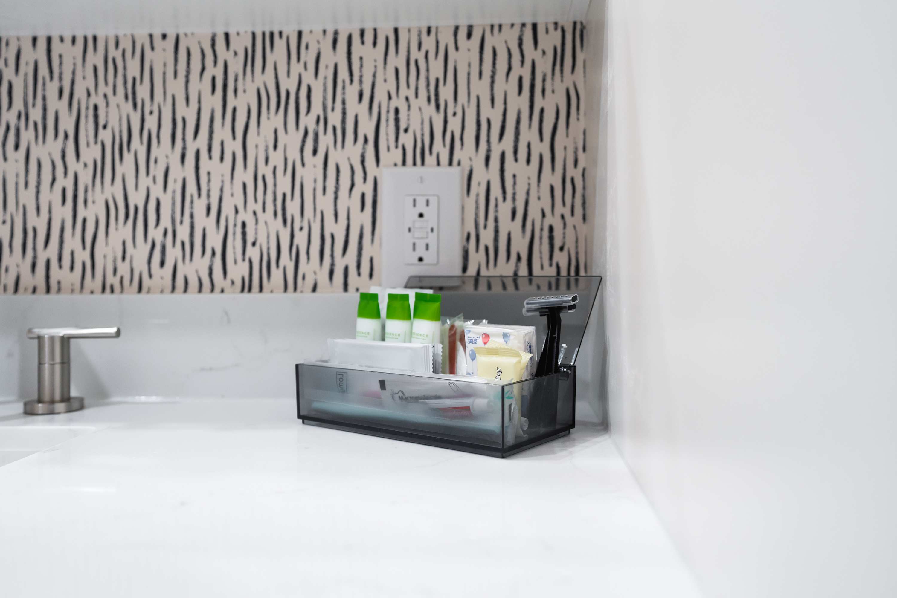 A close-up of a white bathroom countertop shows a clear organizer filled with various toiletries including small shampoo bottles, a razor, and other personal care items. The wall features a textured black and white patterned wallpaper or tile.