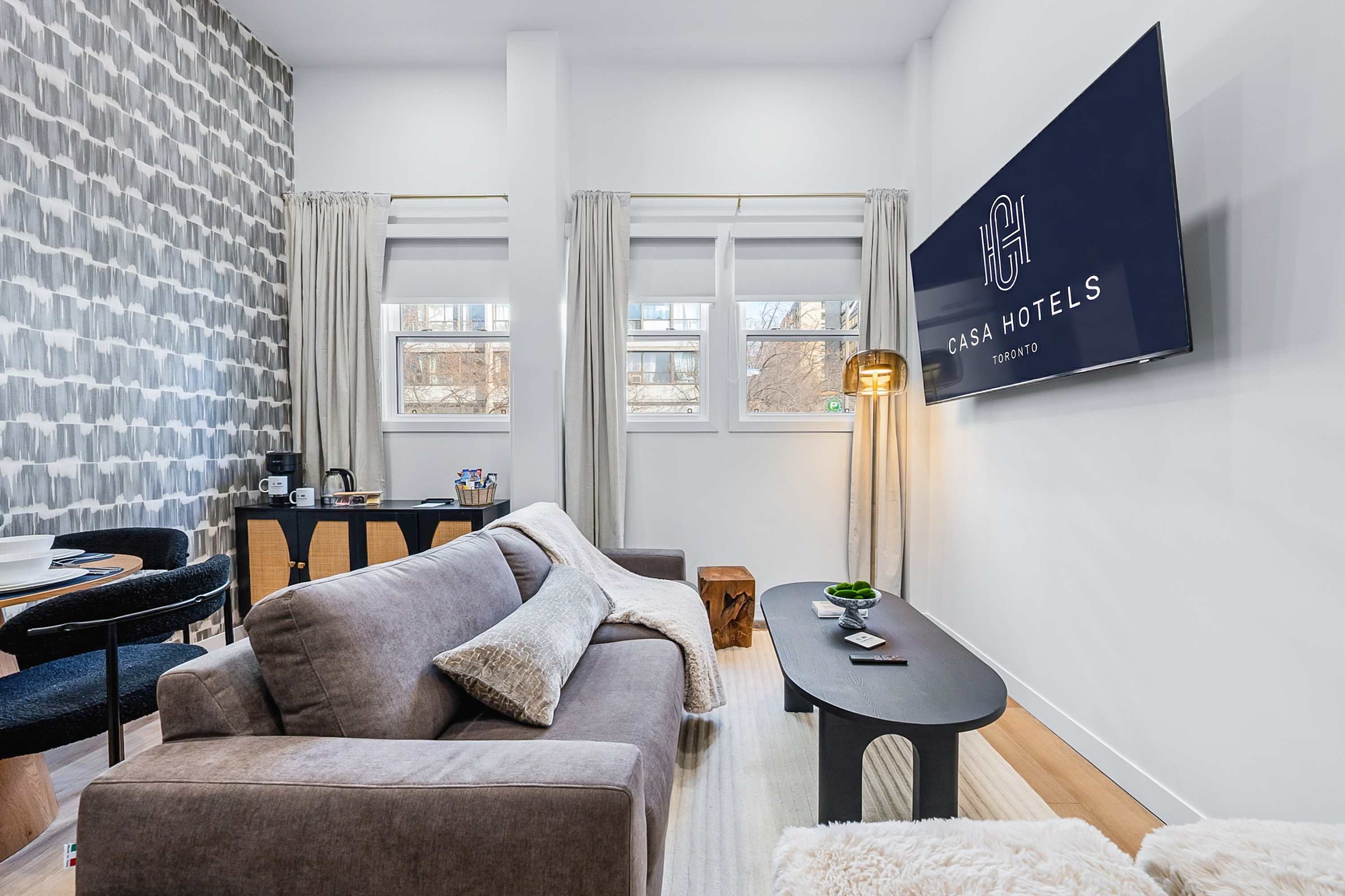 Inviting living room with a comfortable grey sofa adorned with a cream throw blanket and pillows, a dark oval coffee table, and a wall-mounted television displaying the Casa Hotels logo, all illuminated by natural light from multiple windows.