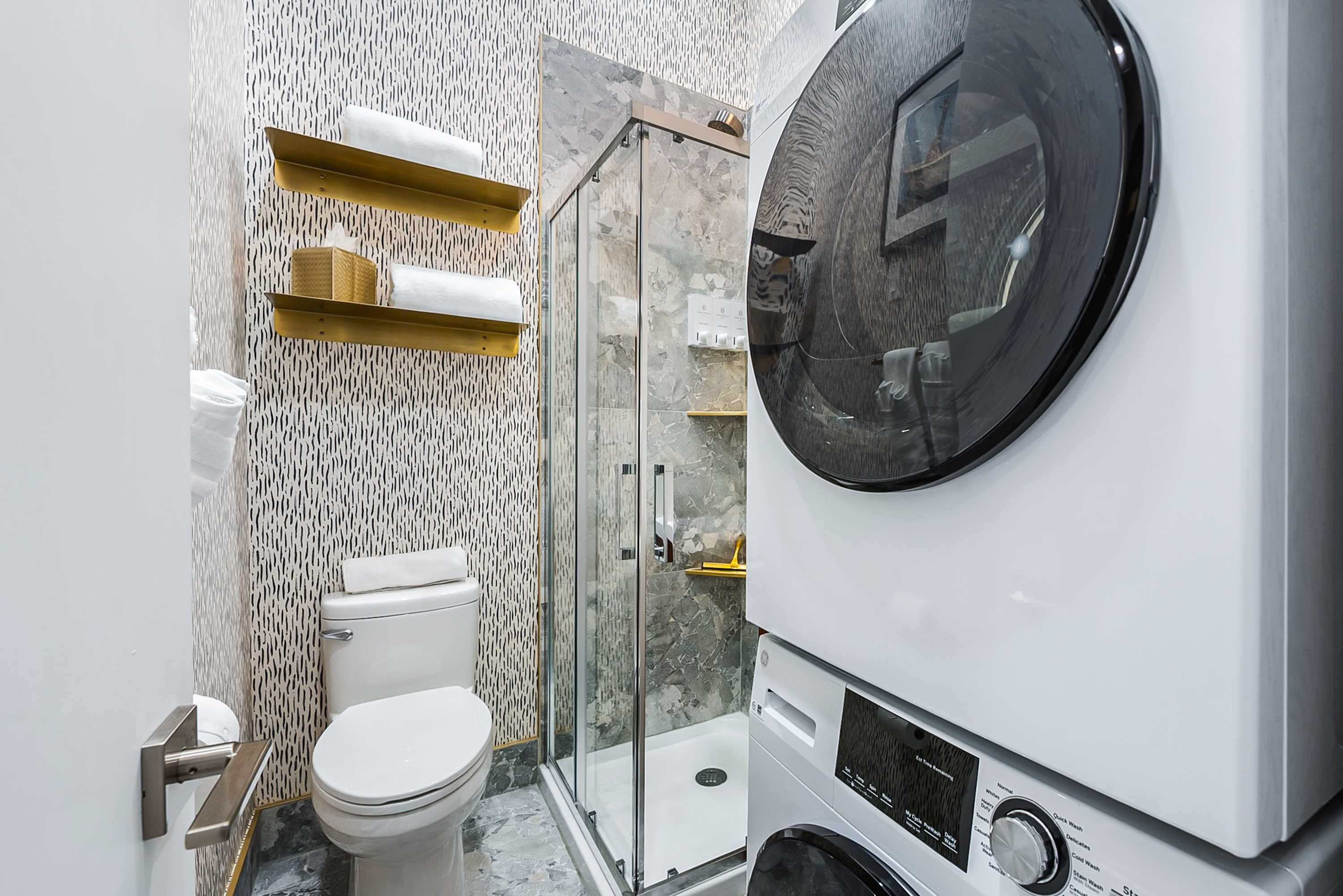 A compact, modern bathroom features a white toilet next to a glass-enclosed shower with grey marble-patterned tiles and gold-toned shelves. A stacked white washer and dryer unit is prominently placed on the right, with patterned wallpaper on the left wall.