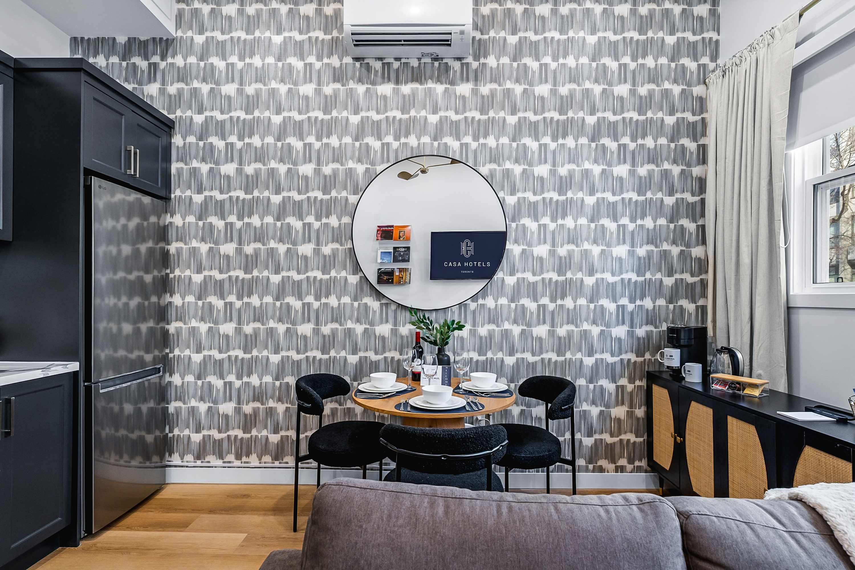 Cozy dining area with a round wooden table and four black chairs, set against a distinctive grey and white patterned wallpaper, with a large round mirror reflecting the space, and a grey sofa visible in the foreground.