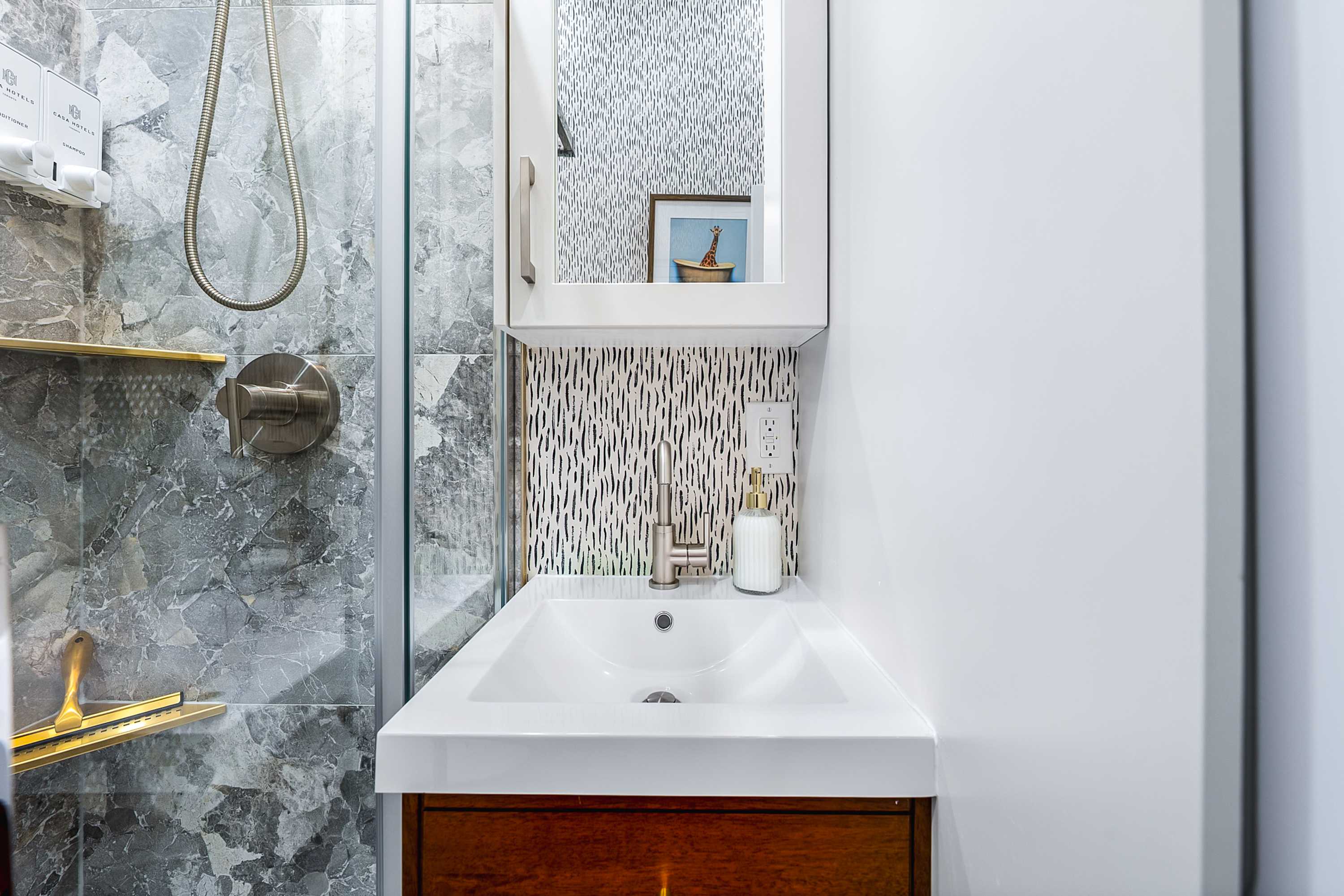 A modern bathroom vanity features a white rectangular sink set into a dark wood cabinet, with a brushed nickel faucet and a mirrored medicine cabinet above. The backsplash is a textured black and white pattern, contrasting with the grey marble-patterned shower tiles.