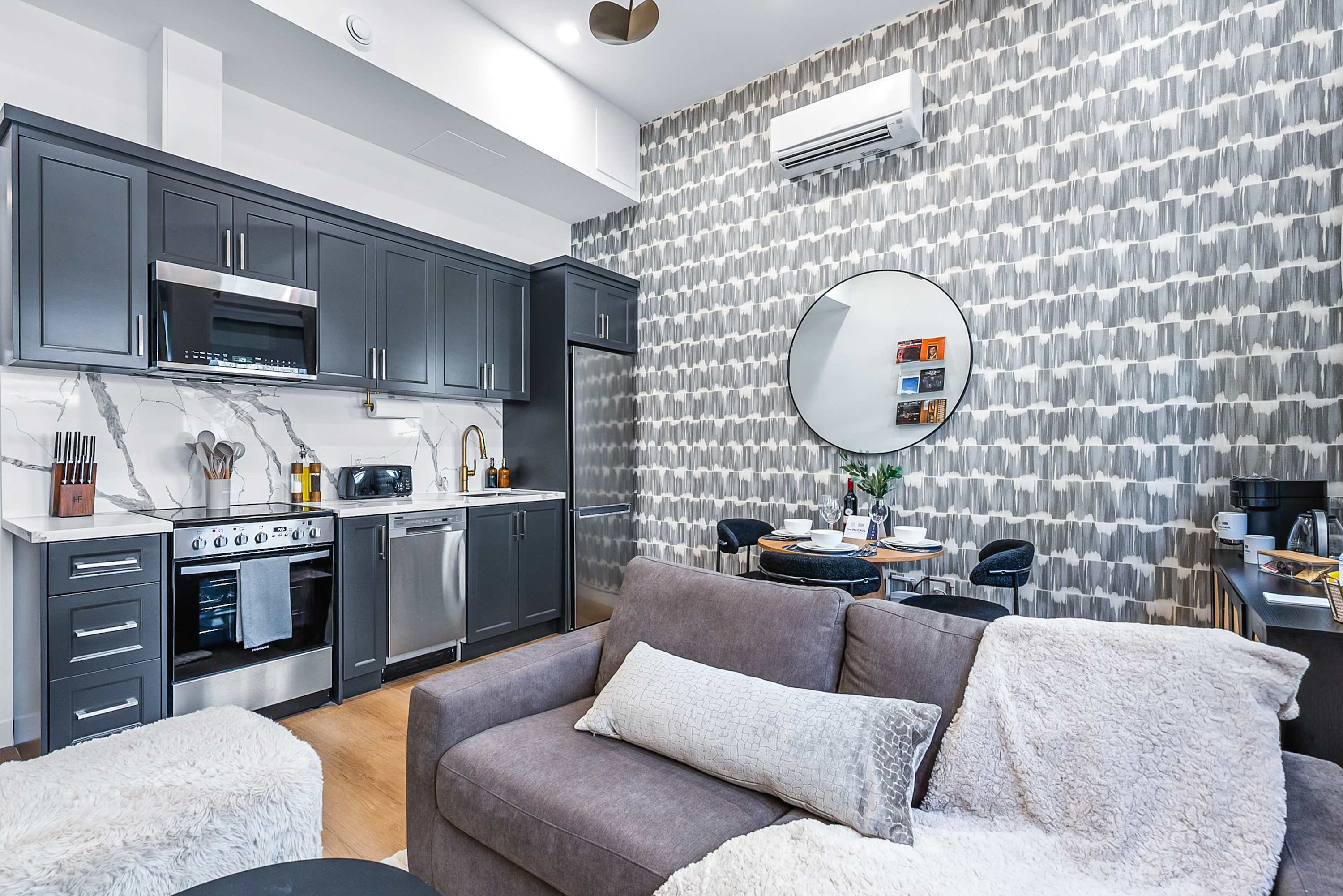 Open-concept living space combining a modern kitchen with dark grey cabinets and stainless steel appliances, a comfortable grey sofa, and a dining area with a round wooden table, all unified by light hardwood floors and a striking patterned accent wall.
