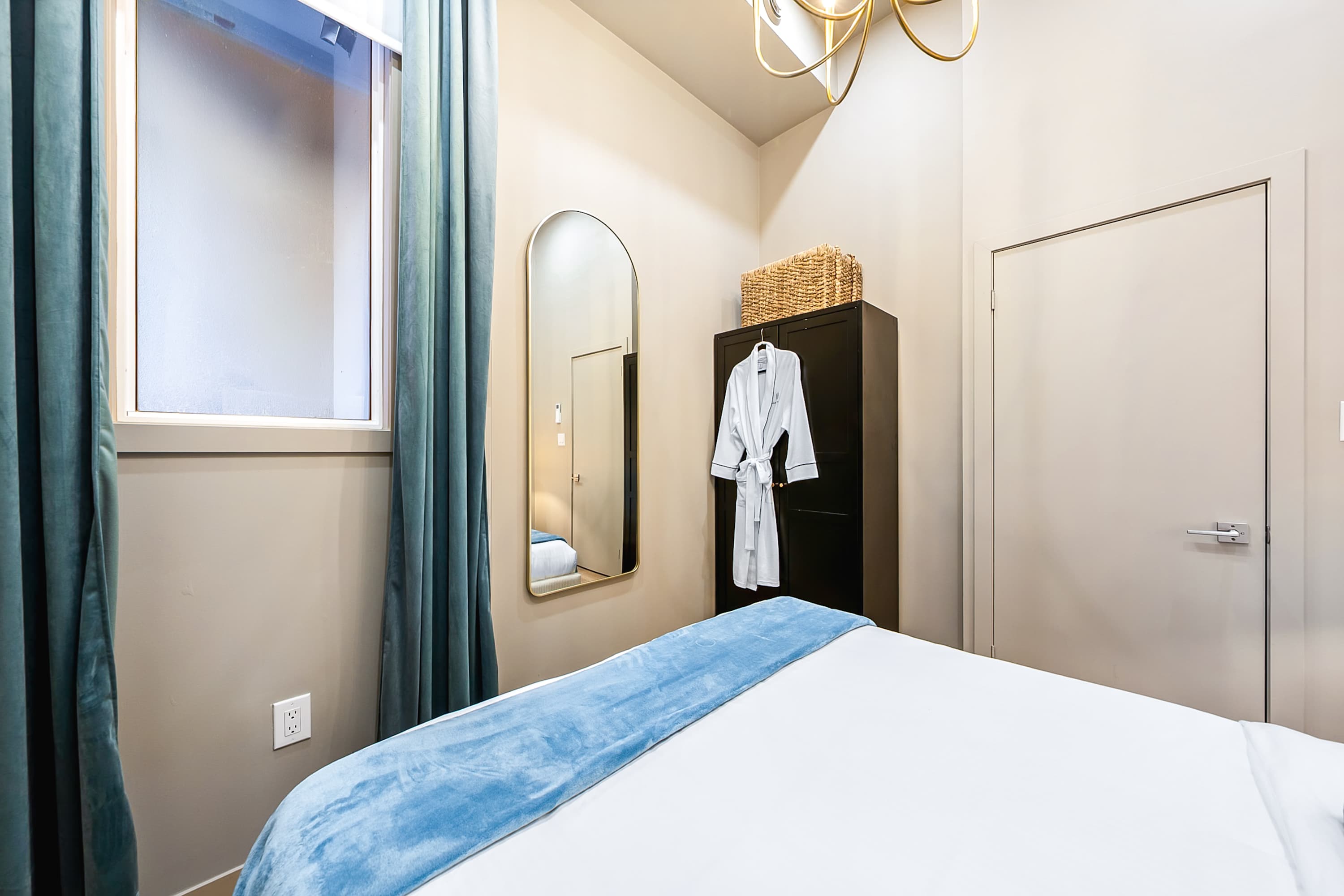 Side view of the bedroom showing a dark wardrobe with a woven basket on top, a full-length arched mirror, and a window with teal curtains, with a white bathrobe hanging on the wardrobe.
