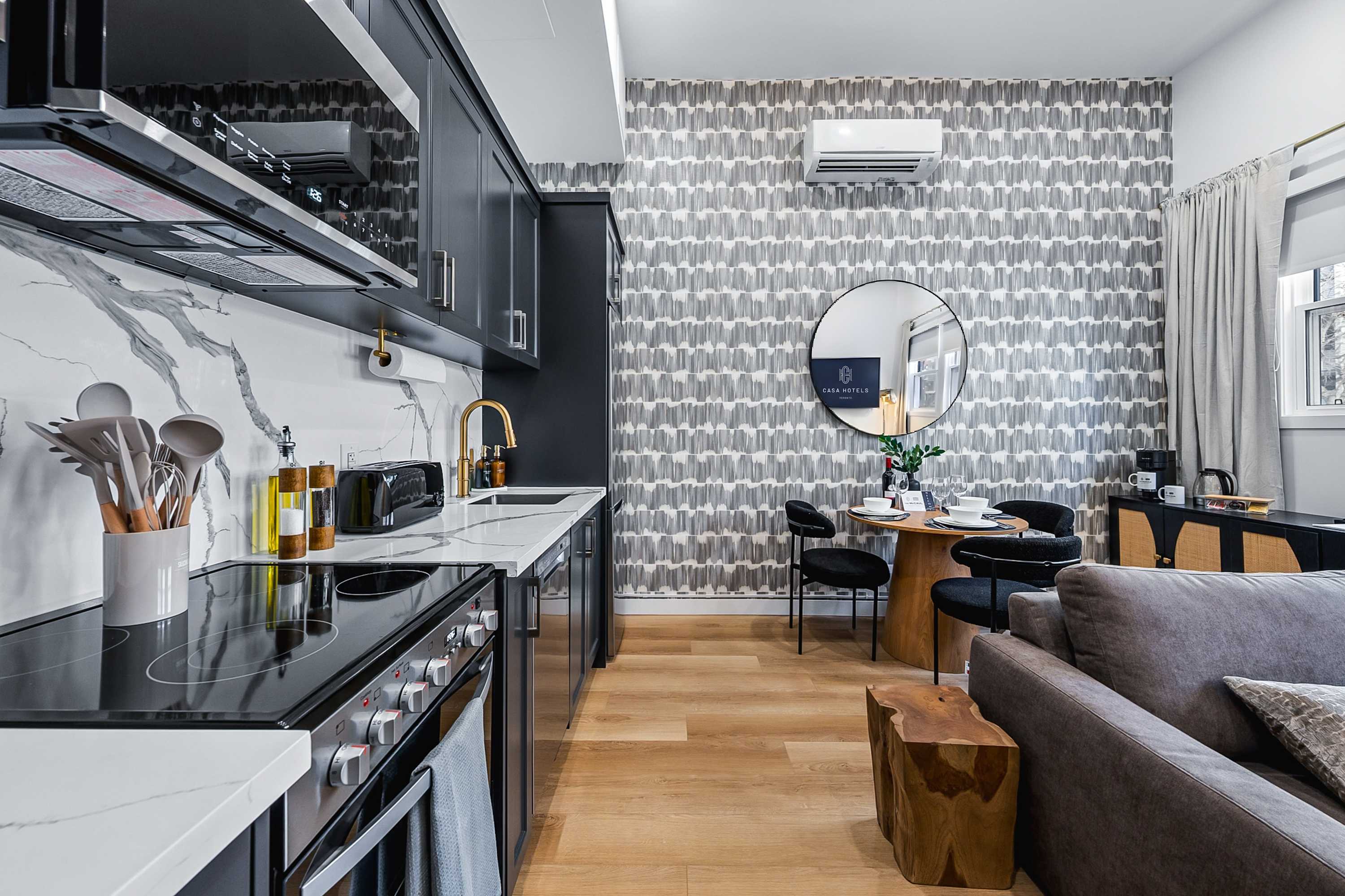 Modern kitchen with dark cabinetry, white marble-patterned backsplash and countertops, and stainless steel appliances, flowing into a dining area with a round wooden table and black chairs, and a glimpse of a living room sofa.