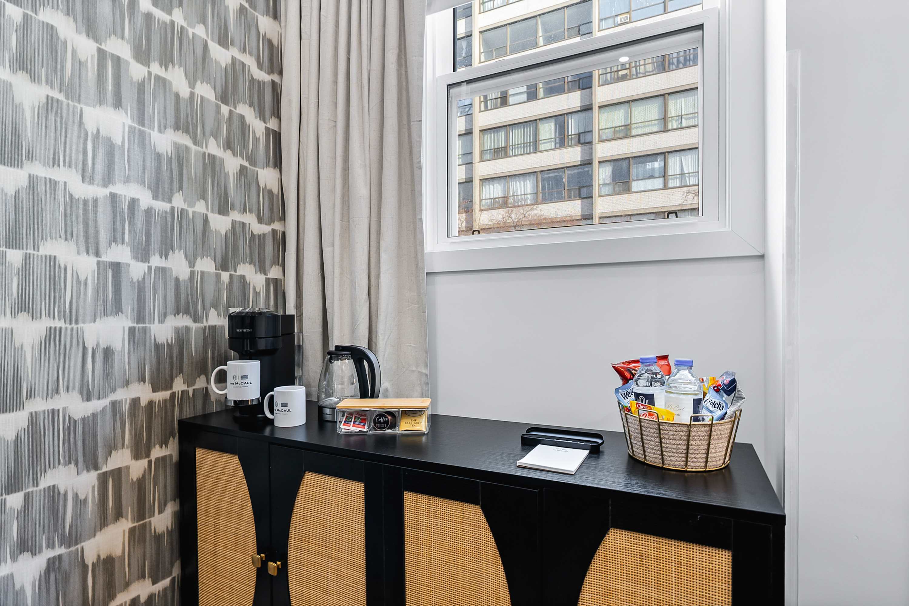 A dedicated coffee and snack station on a black cabinet with rattan accents, featuring a coffee machine, kettle, mugs, and a basket of snacks, positioned next to a window with light-colored curtains.
