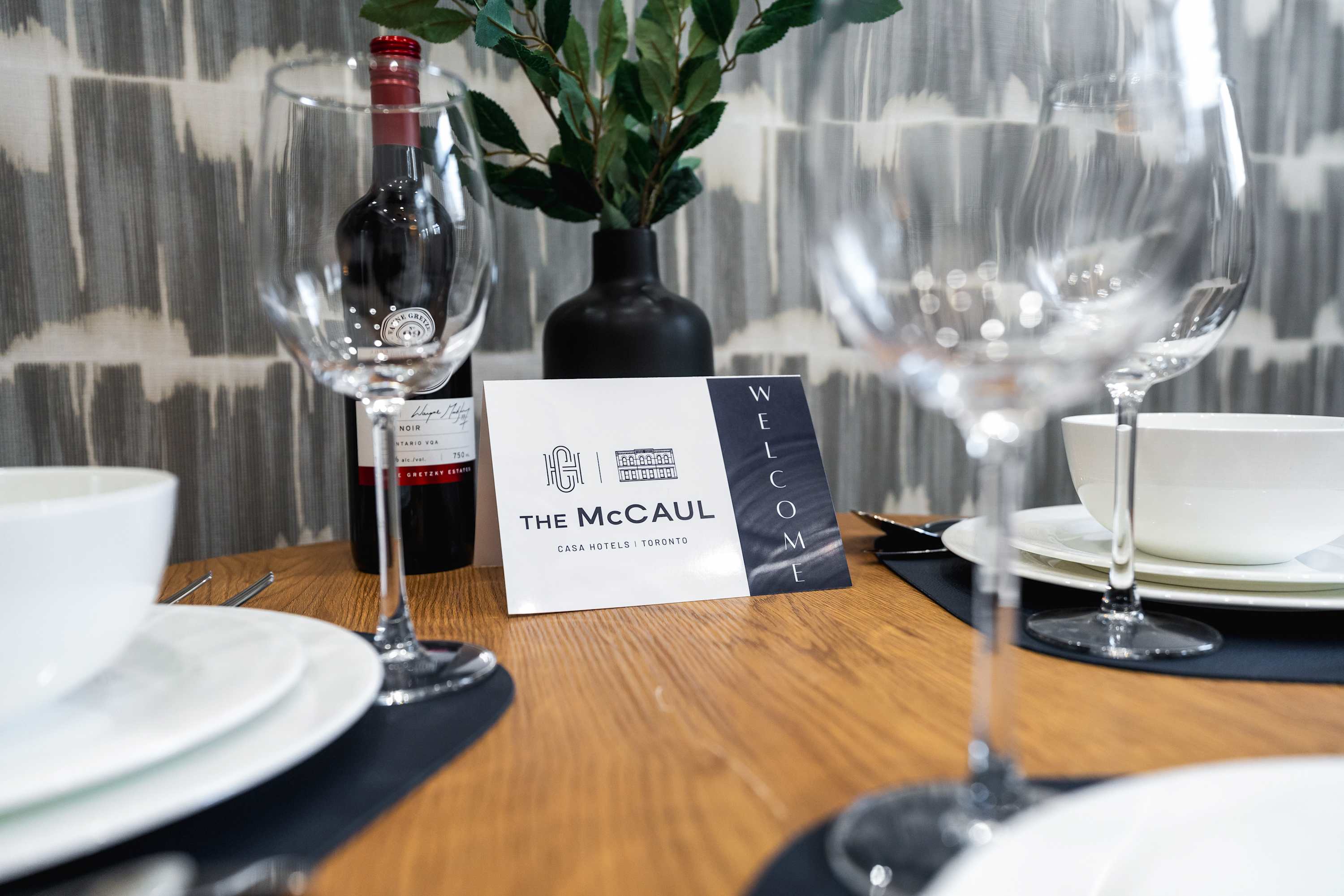 Close-up of a dining table set with white plates, wine glasses, and a bottle of red wine, featuring a "The McCaul" welcome card and a small green plant in a black vase.