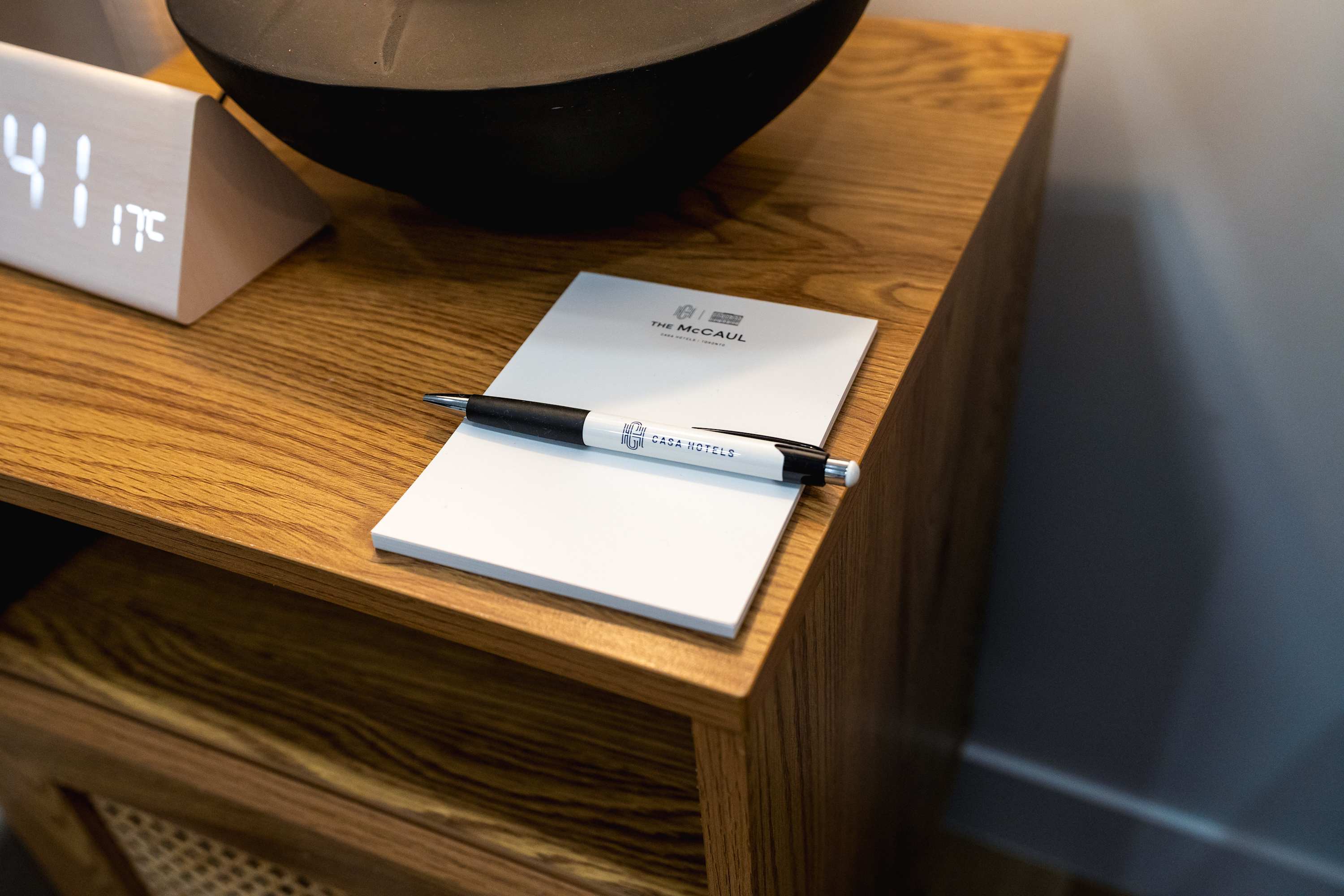 Close-up of a wooden nightstand featuring a digital clock, a black decorative bowl, and hotel stationery including a notepad and a pen with "CASA HOTELS" branding.