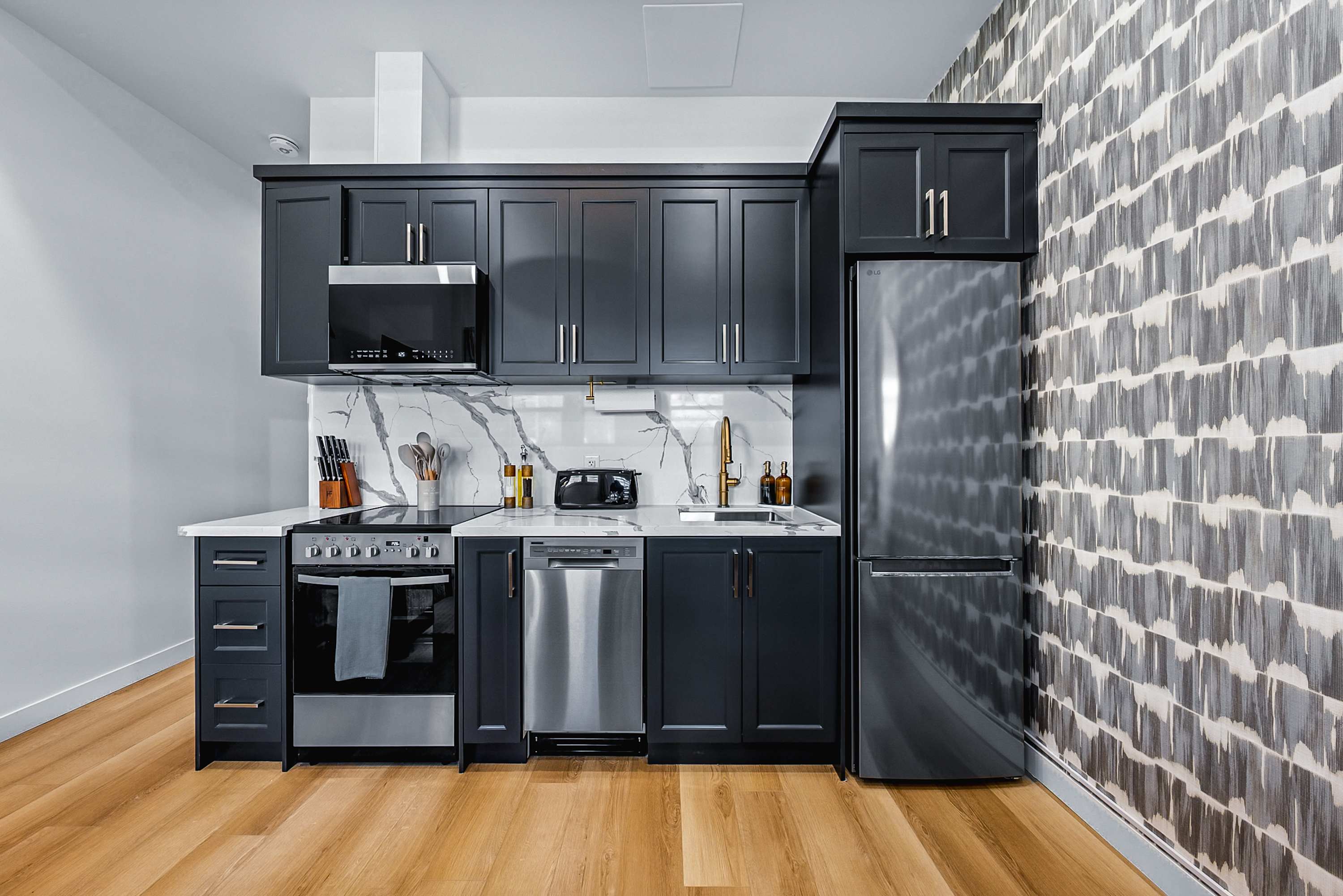 Sleek kitchen featuring dark shaker-style cabinets, white marble-patterned backsplash and countertops, stainless steel appliances including a refrigerator, oven, microwave, and dishwasher, set against a patterned accent wall.